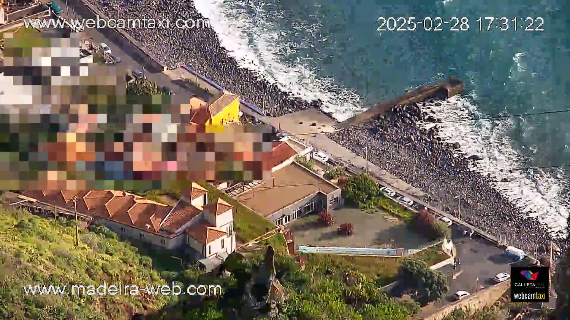 An aerial view shows a rocky coastline with ocean waves, buildings, cars parked along a road, and green vegetation.