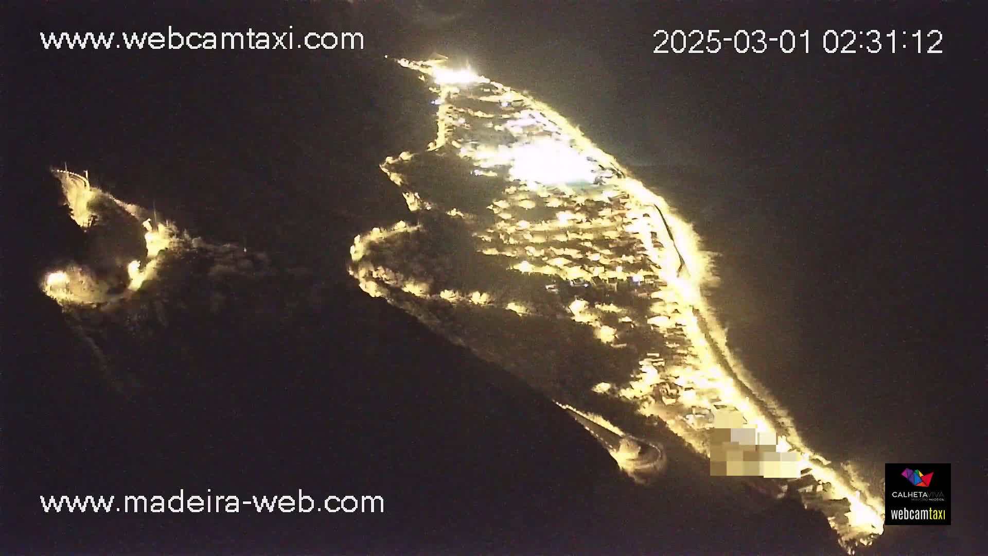 A nighttime aerial view shows a coastal town illuminated by lights along a curving road, nestled against a dark hillside.