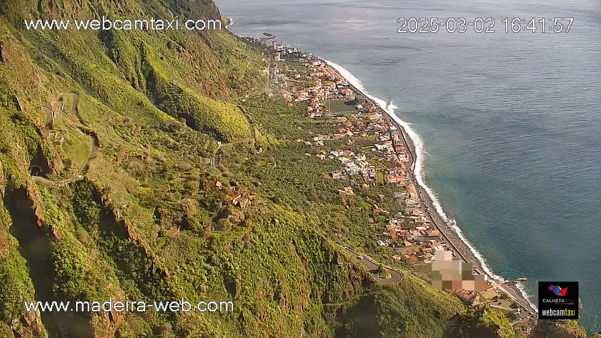 A high-angle, sunny view shows a winding road descending a lush, green cliffside towards a coastal town and dark-blue ocean.
