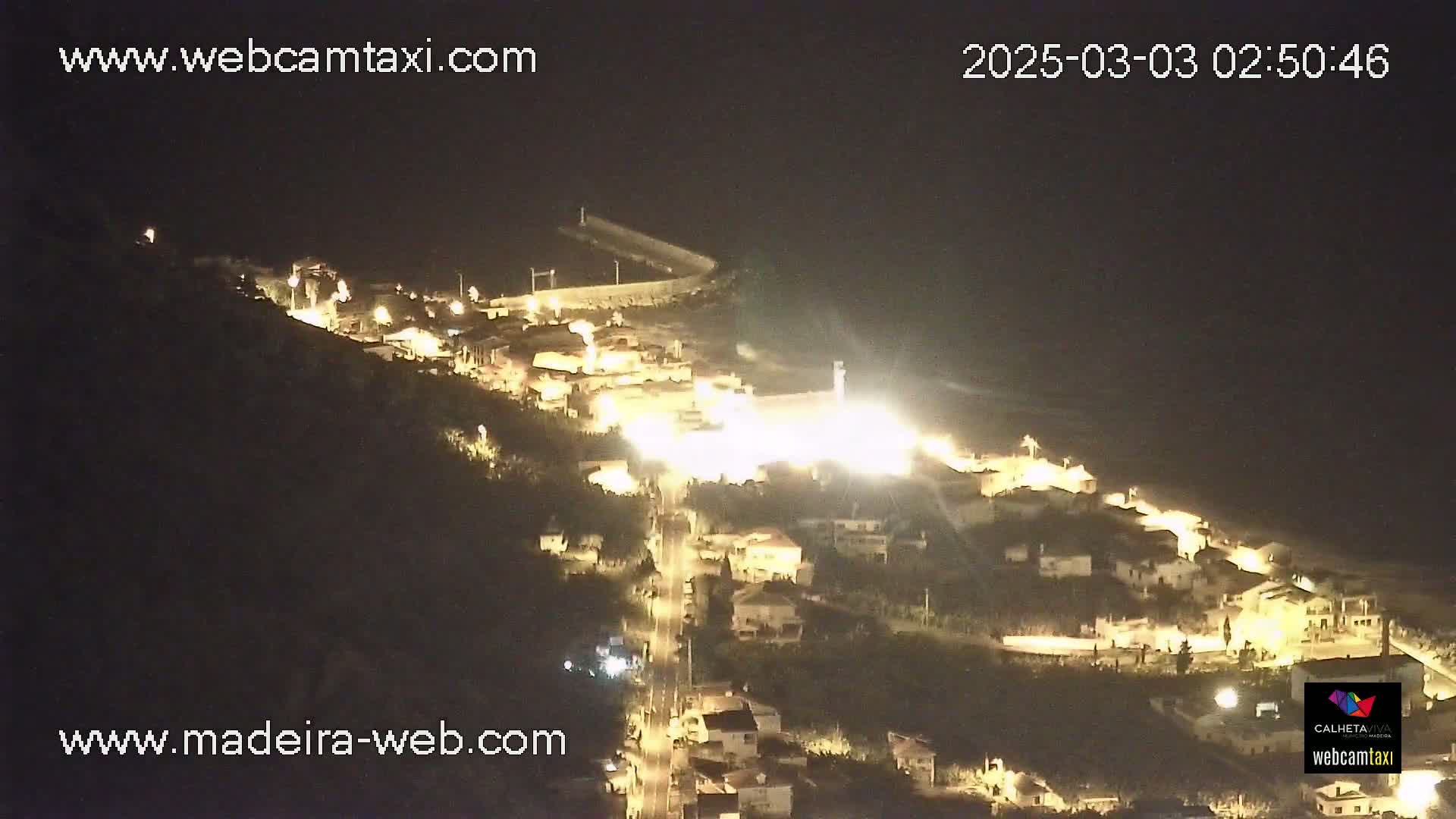 A nighttime aerial view of a coastal town illuminated by streetlights, with a dark body of water visible in the background under a clear sky.