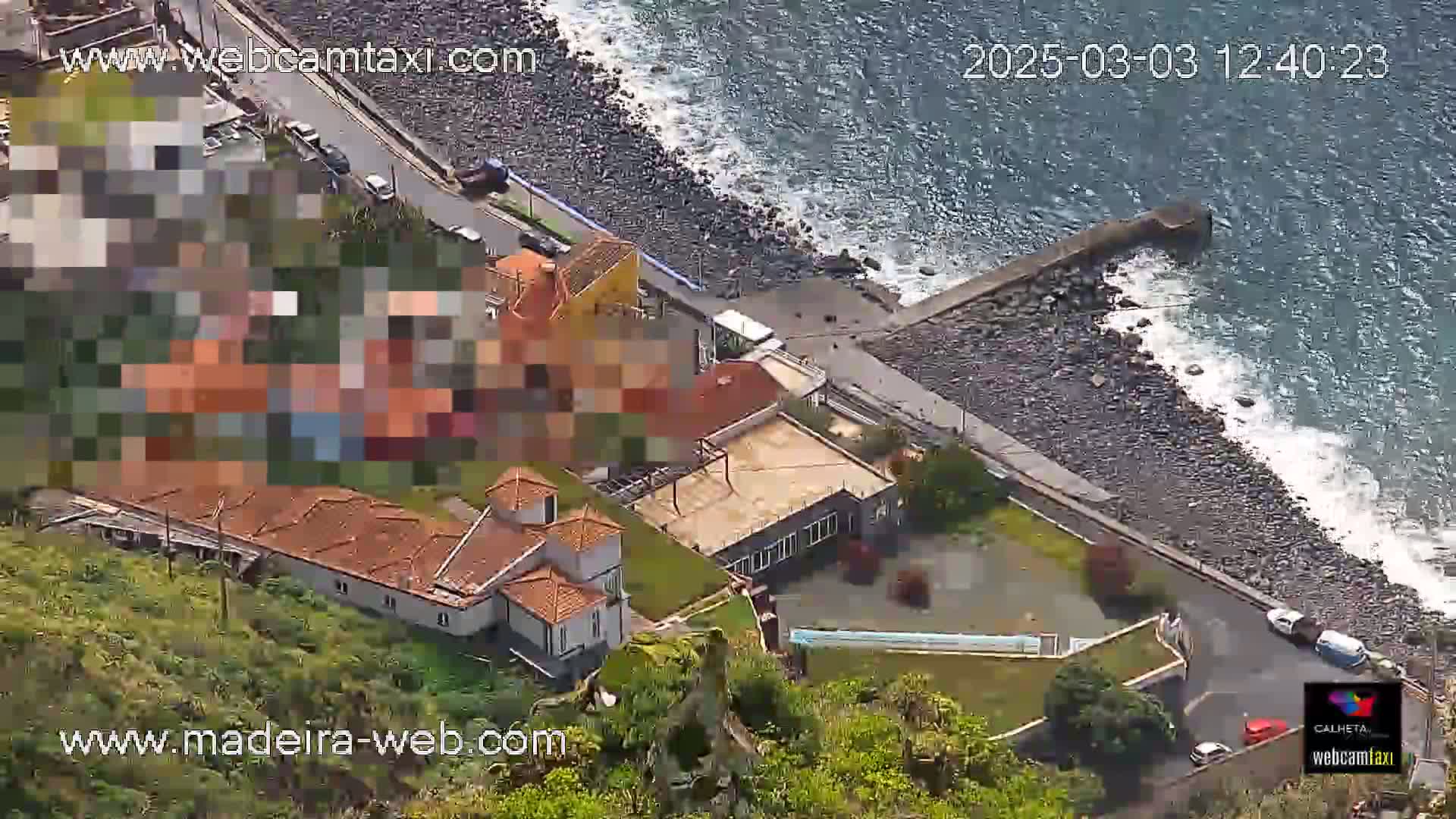 An aerial view shows a rocky coastline with a small village featuring several buildings with red-tiled roofs, next to a calm sea with small waves breaking on the shore.