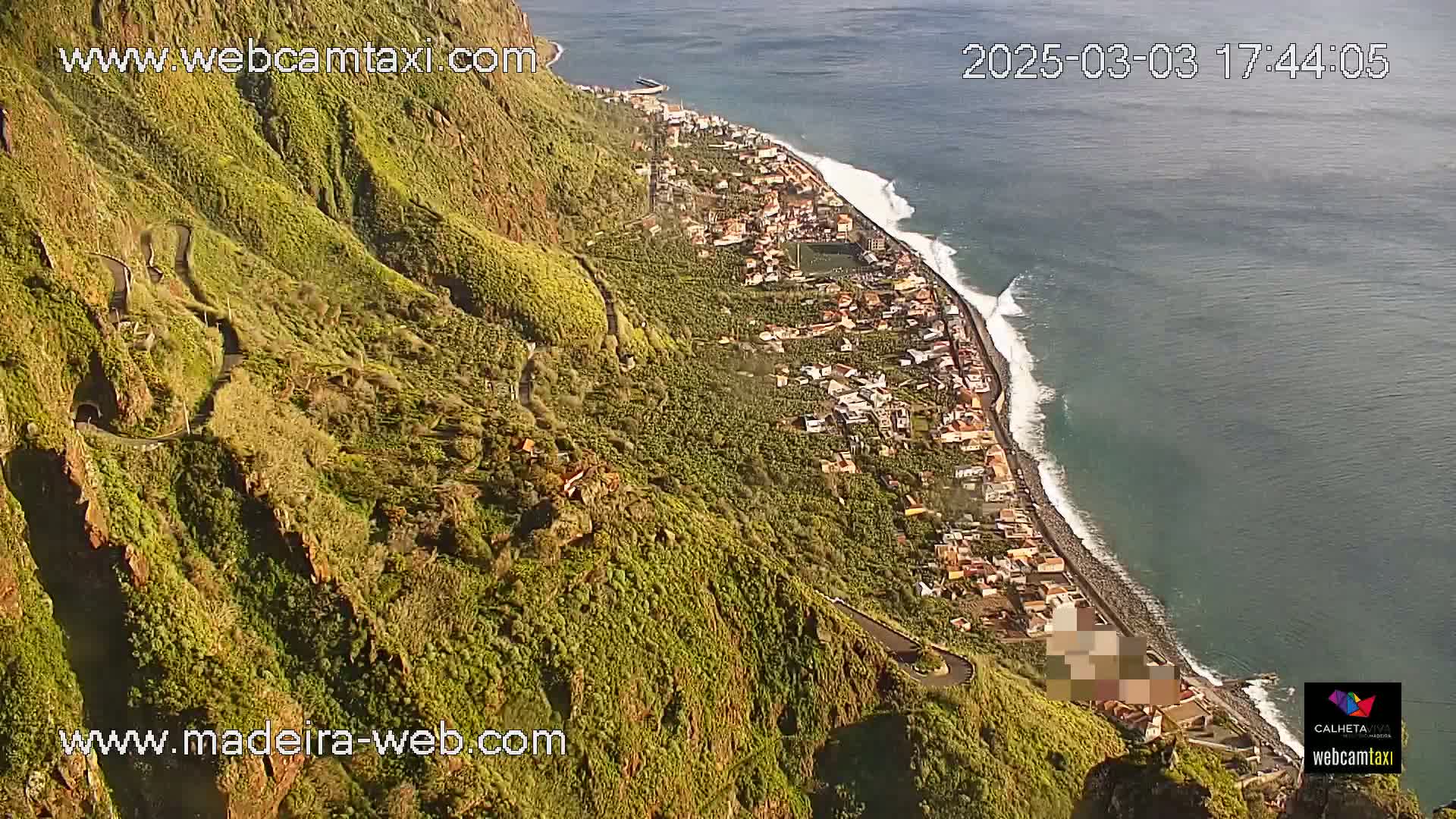 A coastal town nestled along a rocky shoreline with a winding road climbing a steep, green hillside, under sunny conditions.