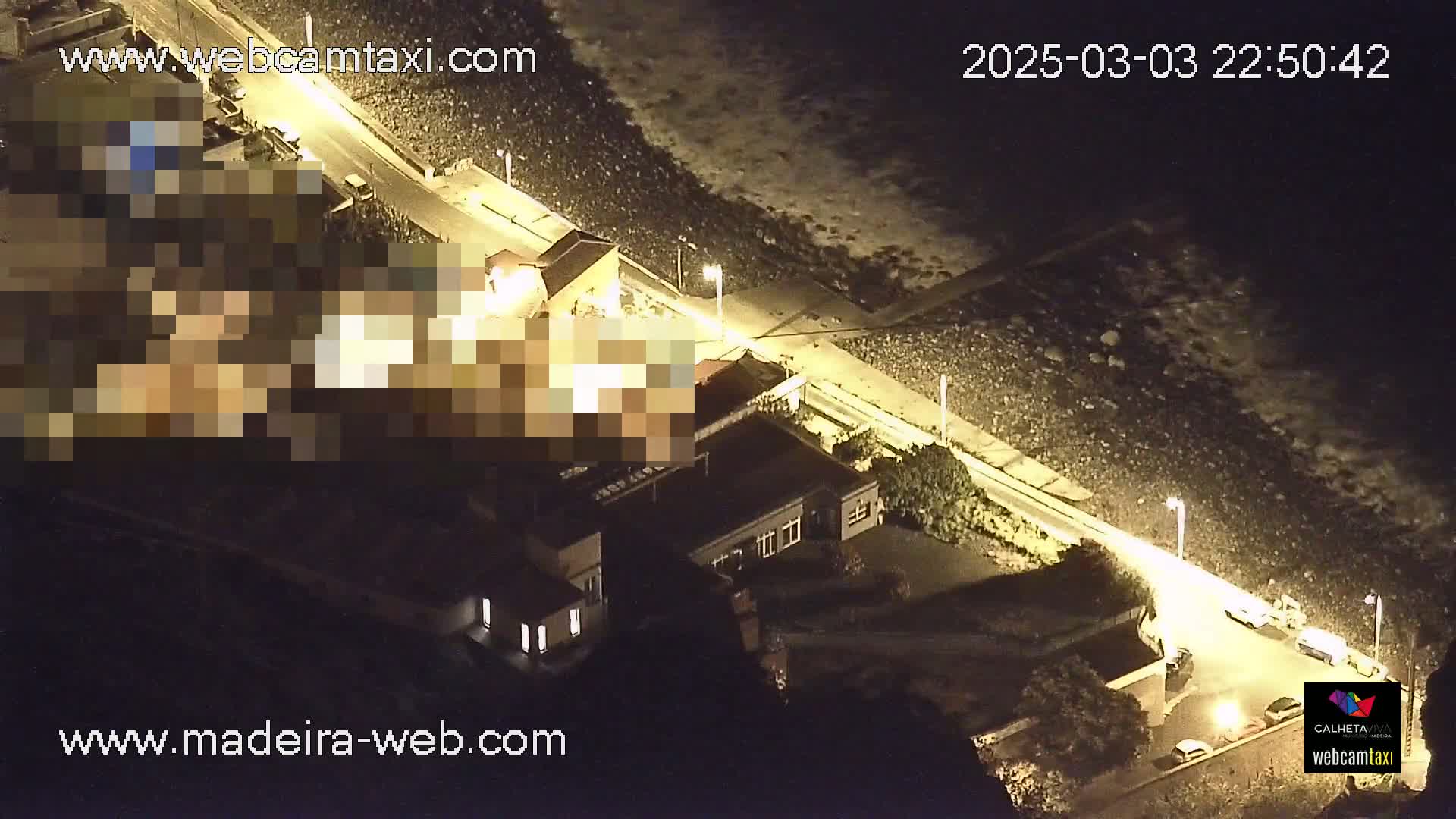 An aerial nighttime view shows a coastal road next to a rocky shoreline with buildings and some parked cars under mostly clear skies.
