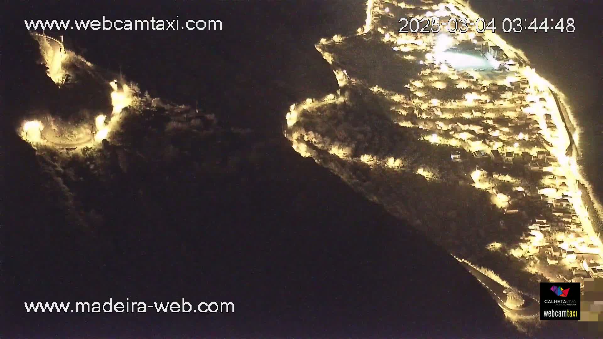An aerial nighttime view shows a coastal town illuminated by lights, situated on a dark hillside next to a winding road.