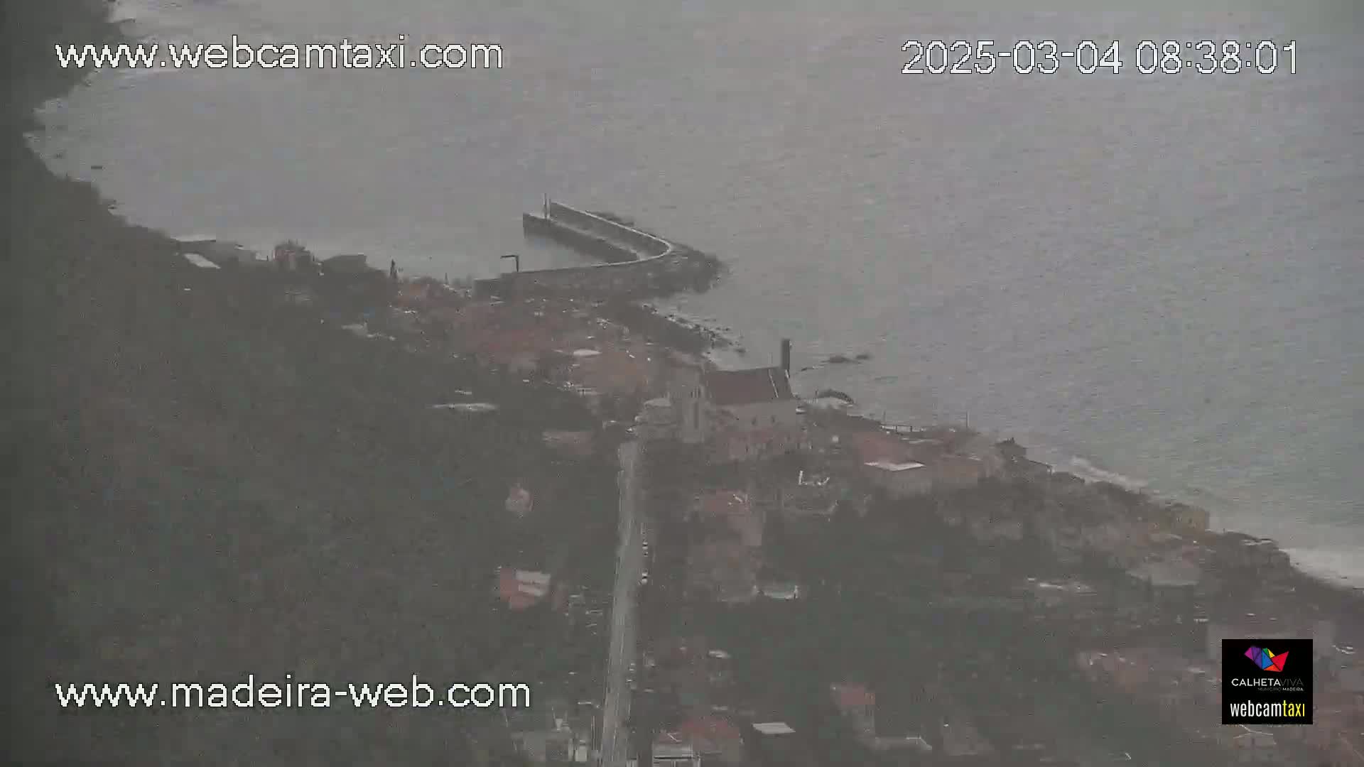 A coastal town is seen from above on an overcast day, with a small harbor and a road winding through the buildings down to the water.