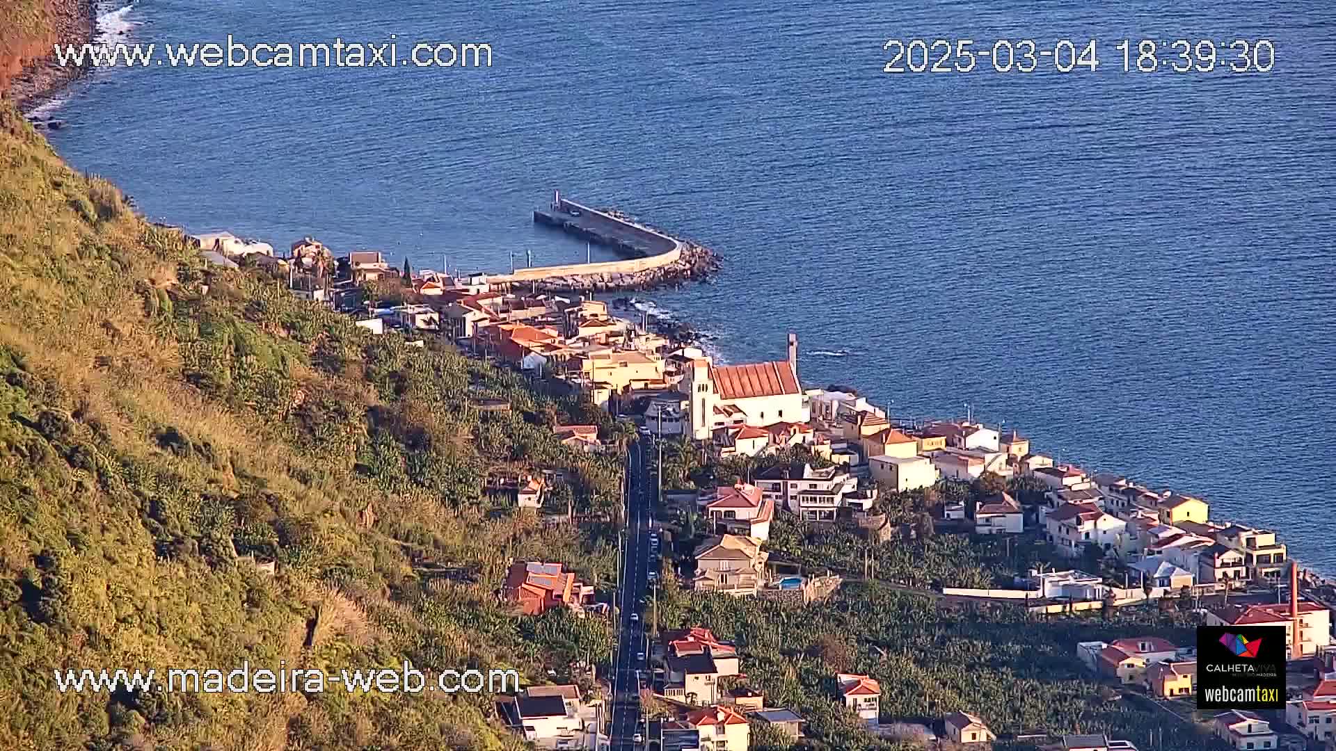 An aerial view shows a coastal town nestled on a hillside overlooking a calm ocean on a sunny day, with a small harbor visible in the foreground.
