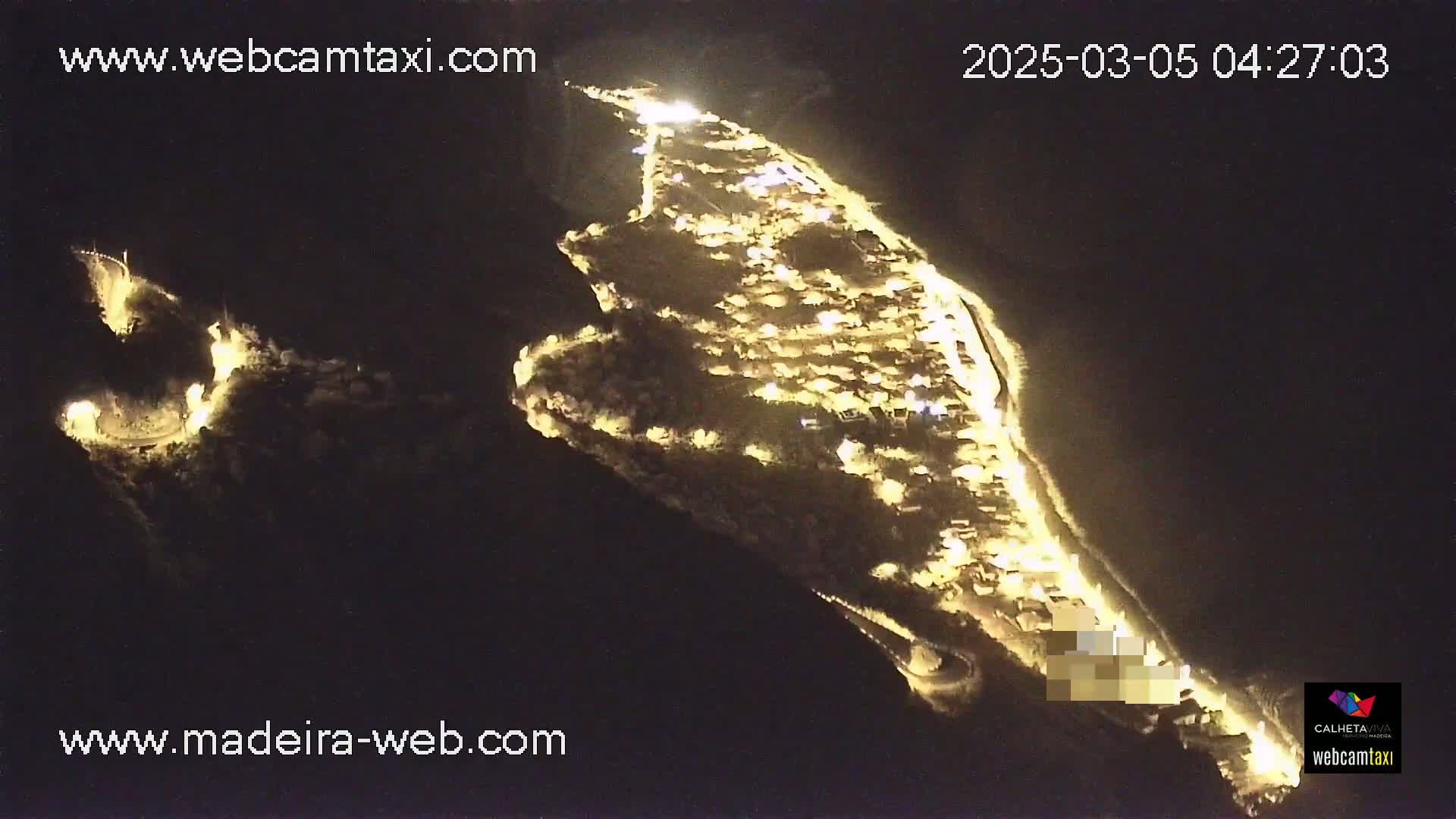 An aerial nighttime view shows a coastal town brightly lit up along a dark, curving coastline.