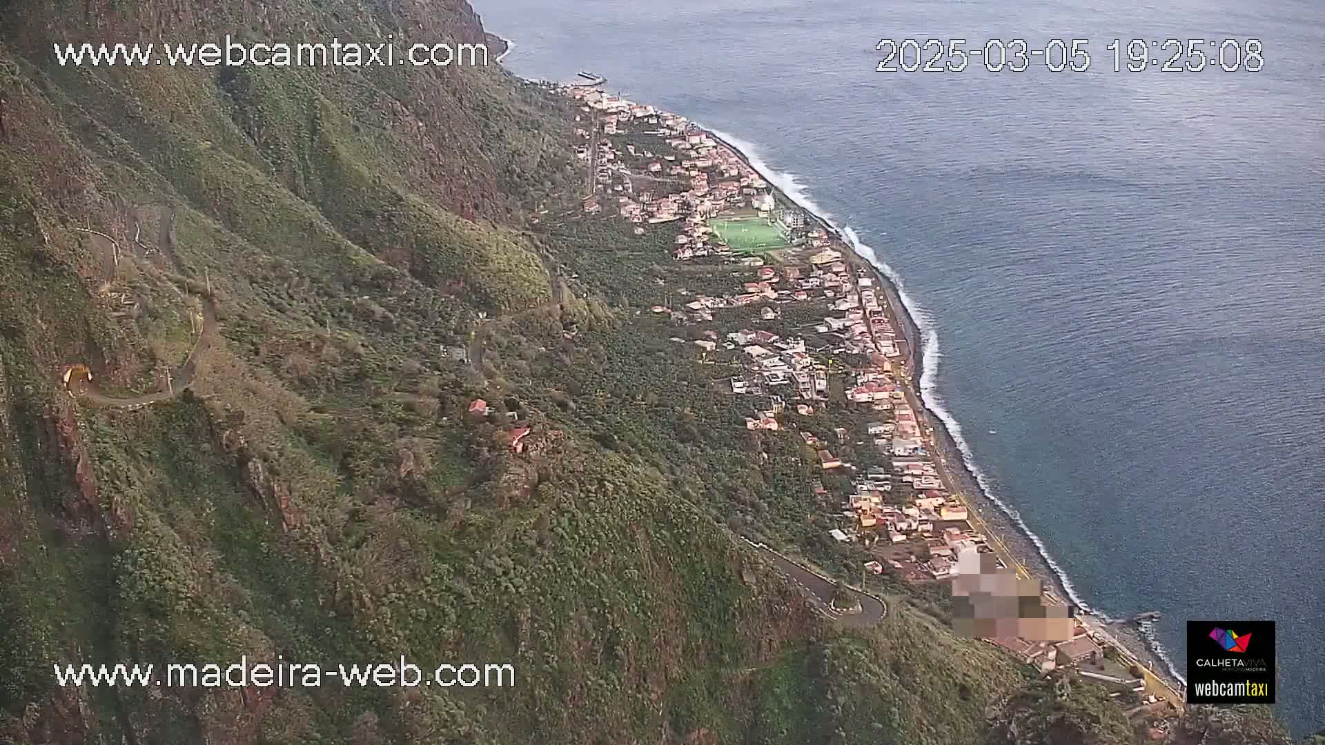 An aerial view shows a coastal town nestled between a steep, green mountain and a dark-blue ocean under overcast skies.