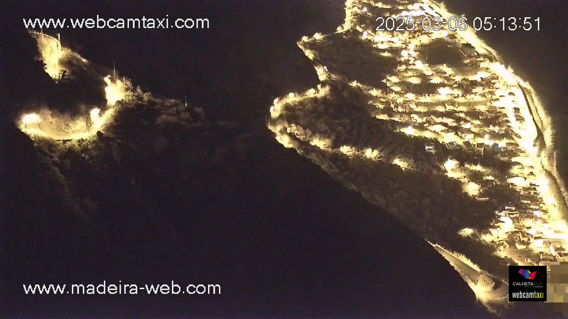 An aerial nighttime view shows a village illuminated by lights nestled on a hillside, with a winding road also lit up on a nearby mountain.