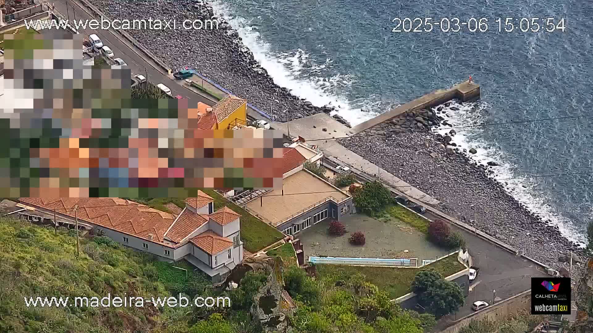An aerial view shows a rocky coastline with moderate waves, a small building complex, and a paved road with parked cars.