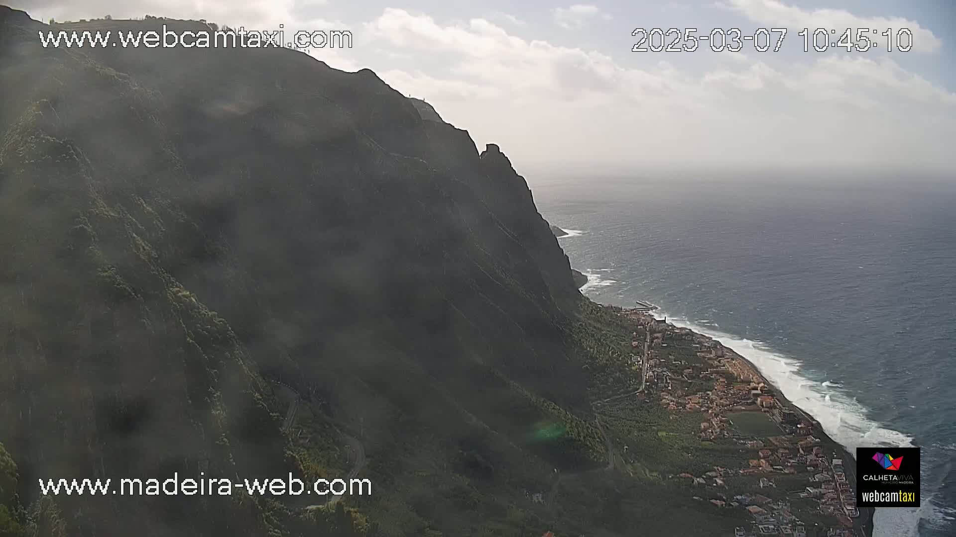 A steep, dark green cliffside overlooks a coastal town and a wavy ocean under a mostly cloudy sky.