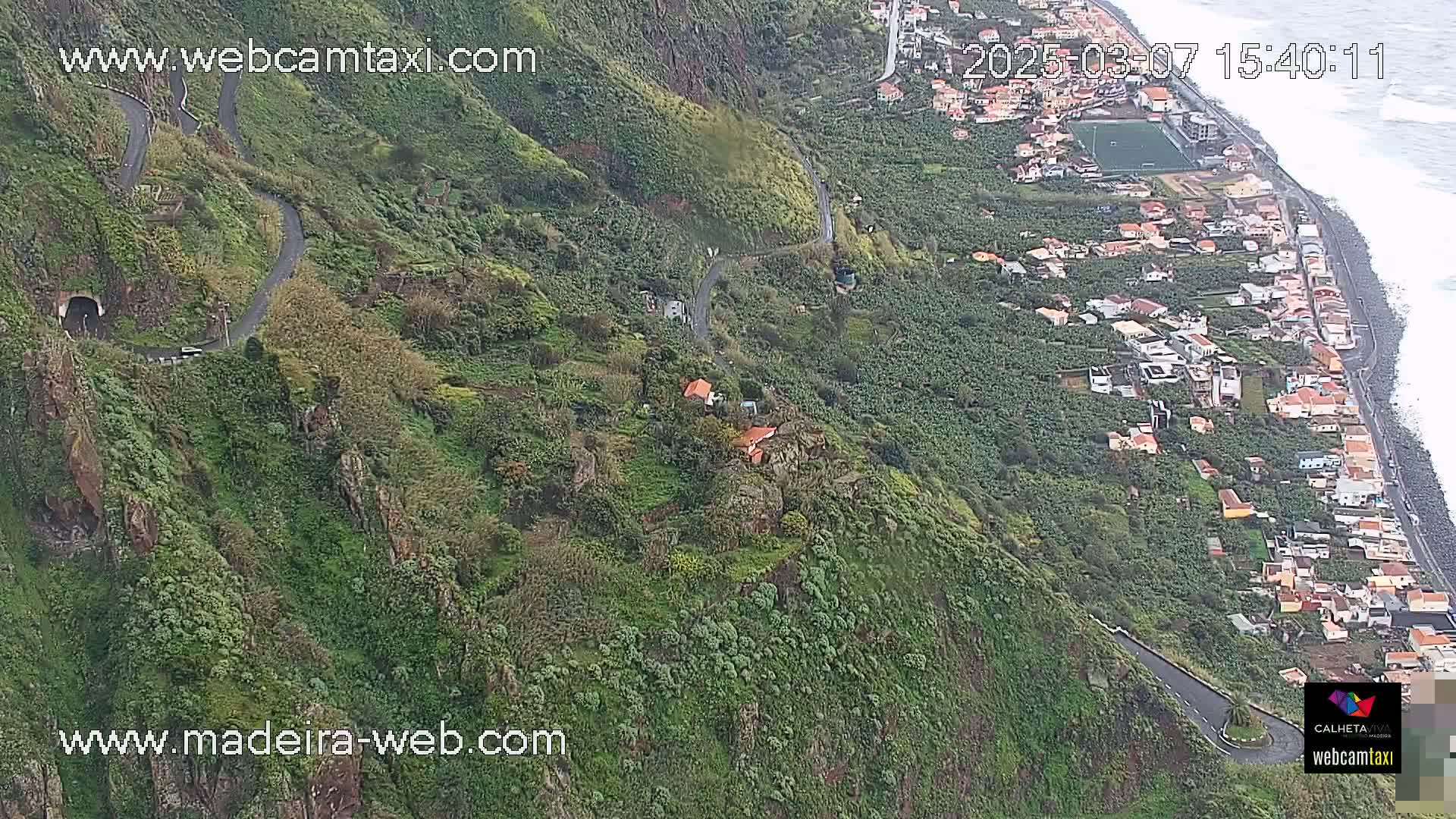 A winding road snakes down a lush, green hillside toward a coastal town with a rocky shoreline and ocean under partly cloudy skies.