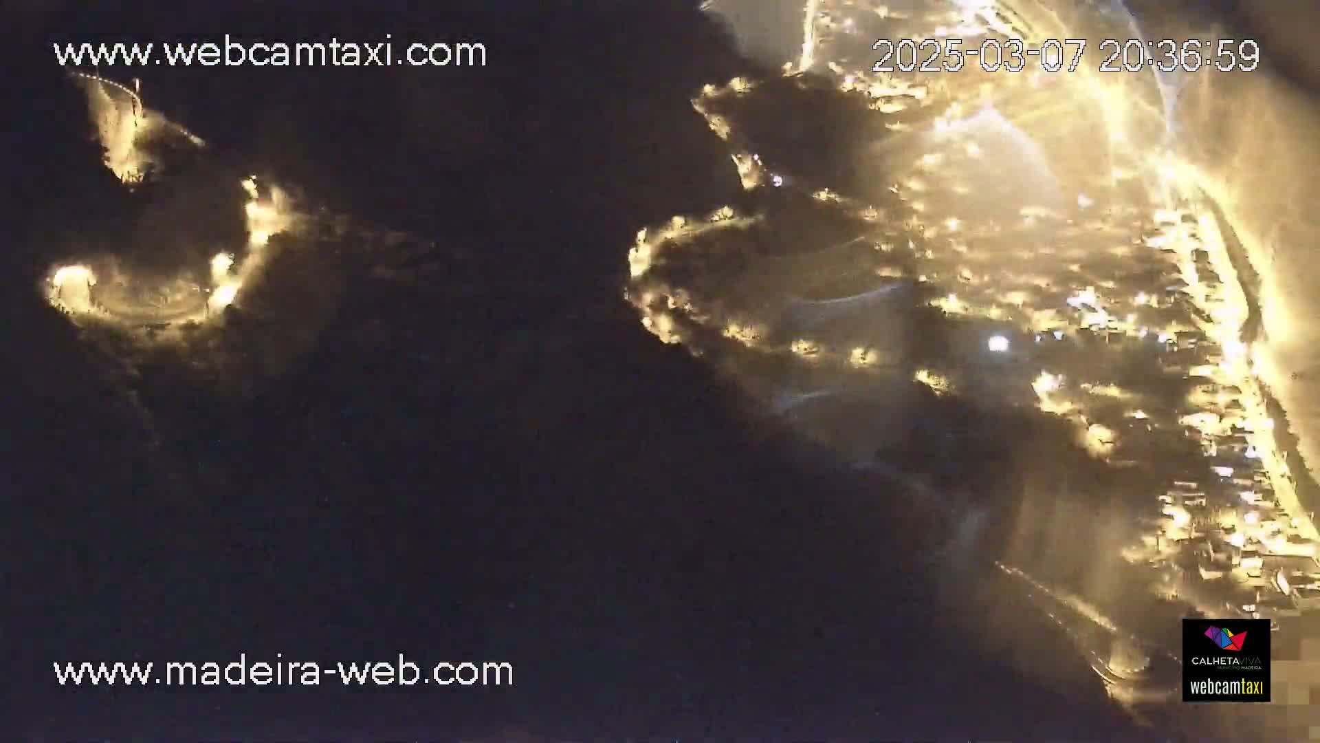 A nighttime aerial view shows a winding road illuminated by lights snaking along a dark hillside towards a brightly lit coastal town.