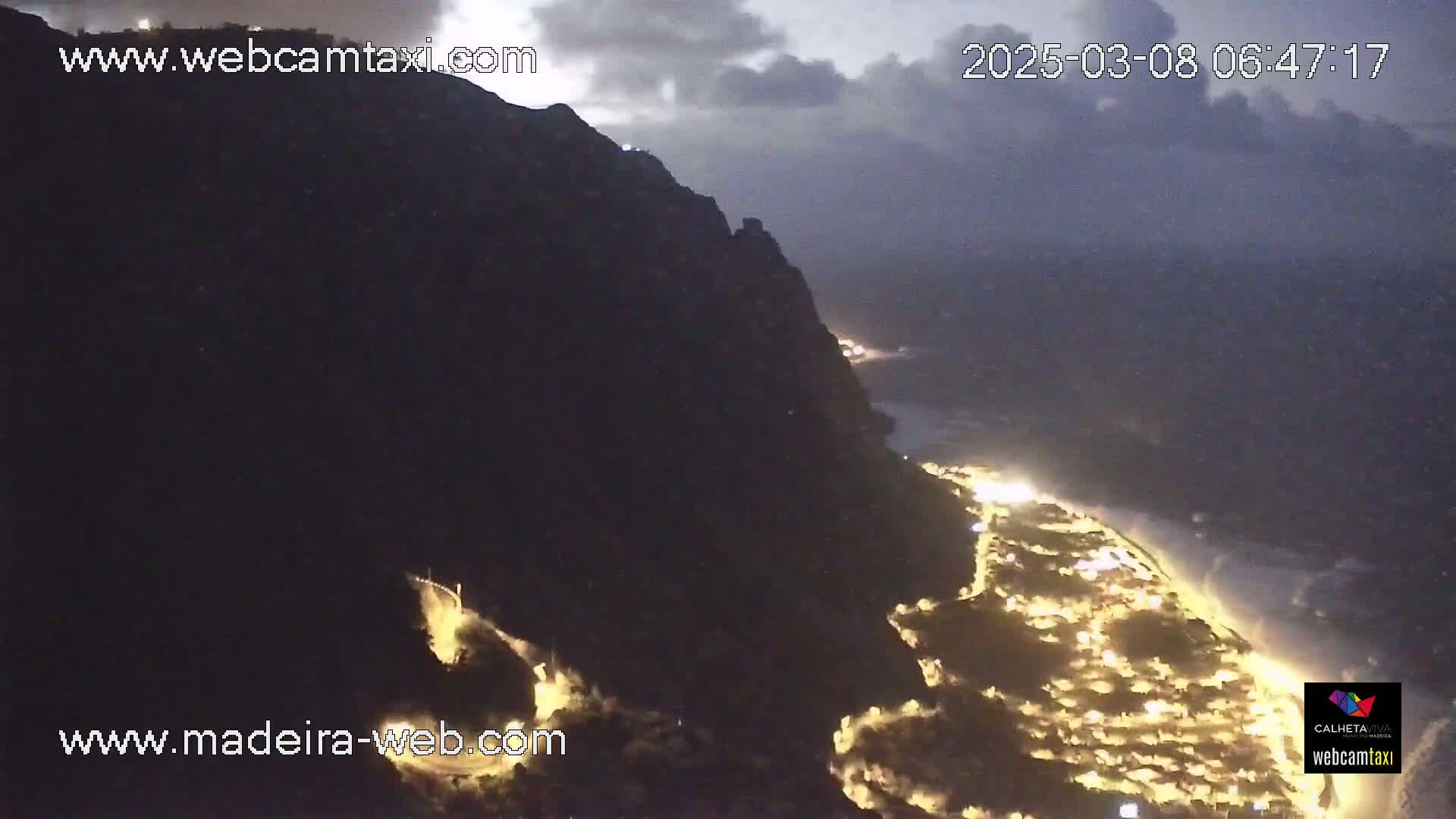 A nighttime, high-angle view shows a dark cliff overlooking a coastal town illuminated by lights, with a dark, cloudy sky.
