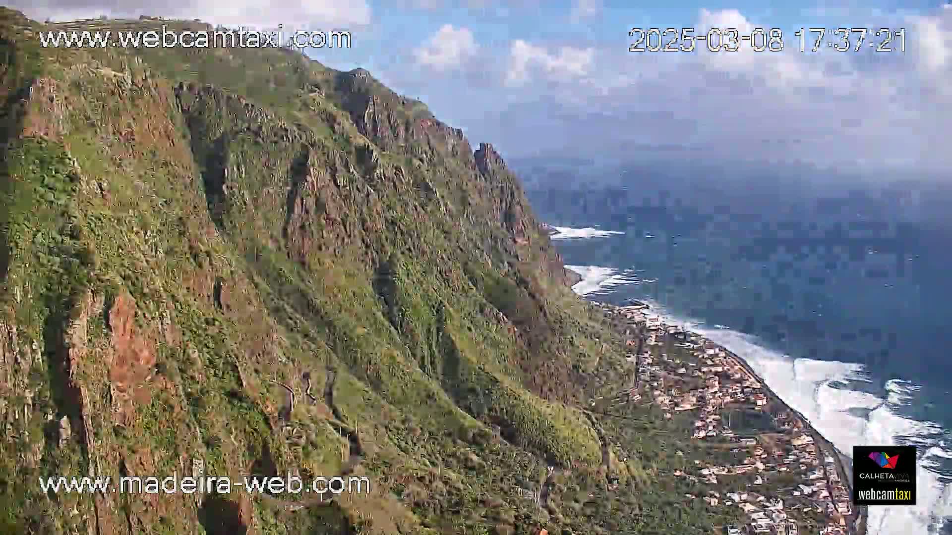 A high-angle view shows a steep, verdant cliffside overlooking a town nestled along a coastline with crashing waves under a partly cloudy sky.