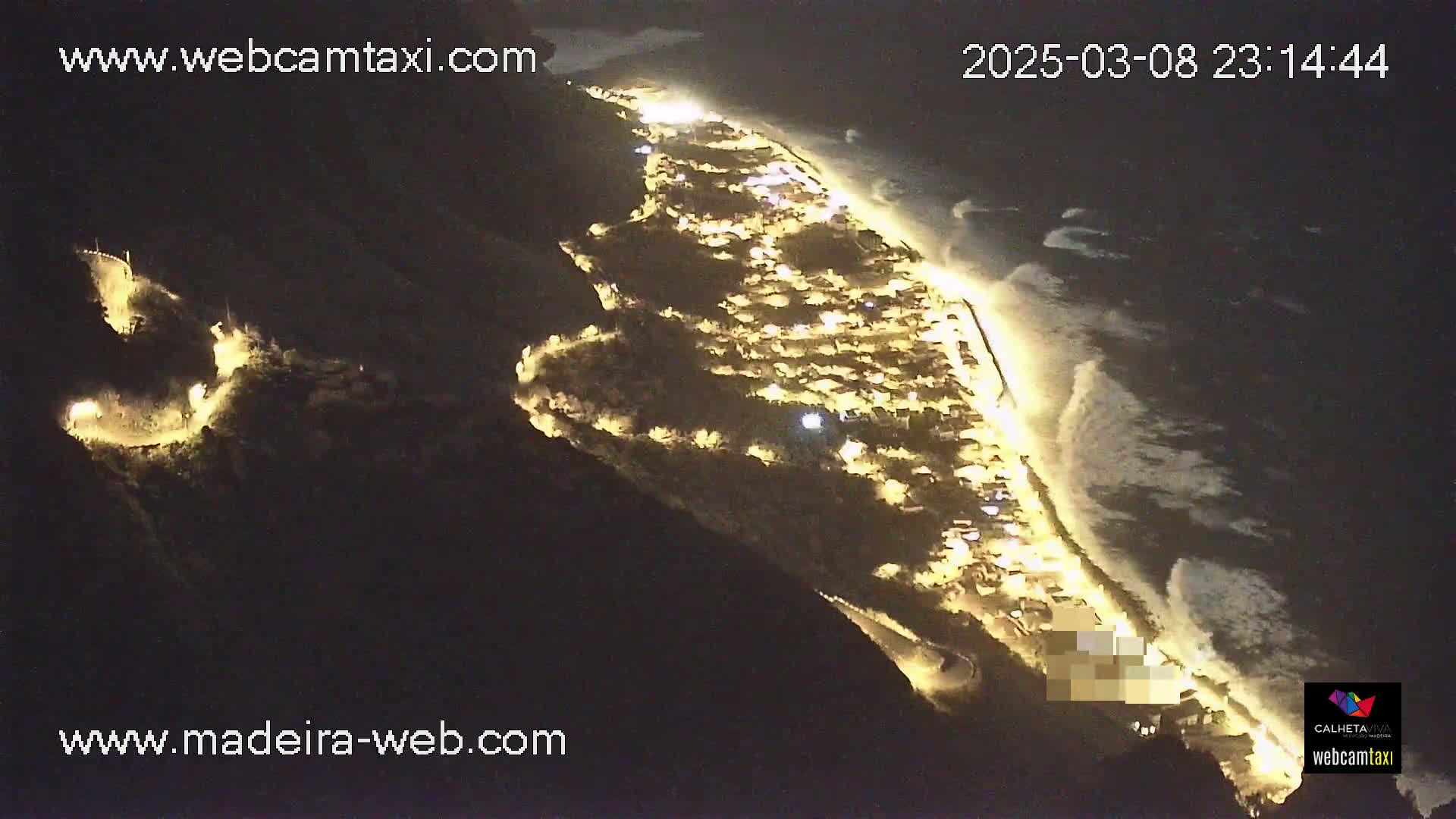A nighttime aerial view of a coastal town illuminated with lights, alongside a dark ocean with waves crashing on the shore.