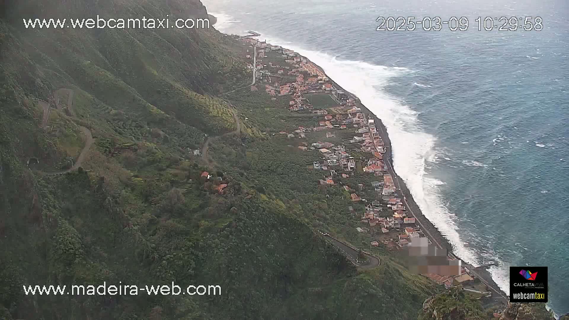 An aerial view shows a coastal town nestled on a hillside overlooking a dark-blue ocean with substantial waves under somewhat overcast skies.
