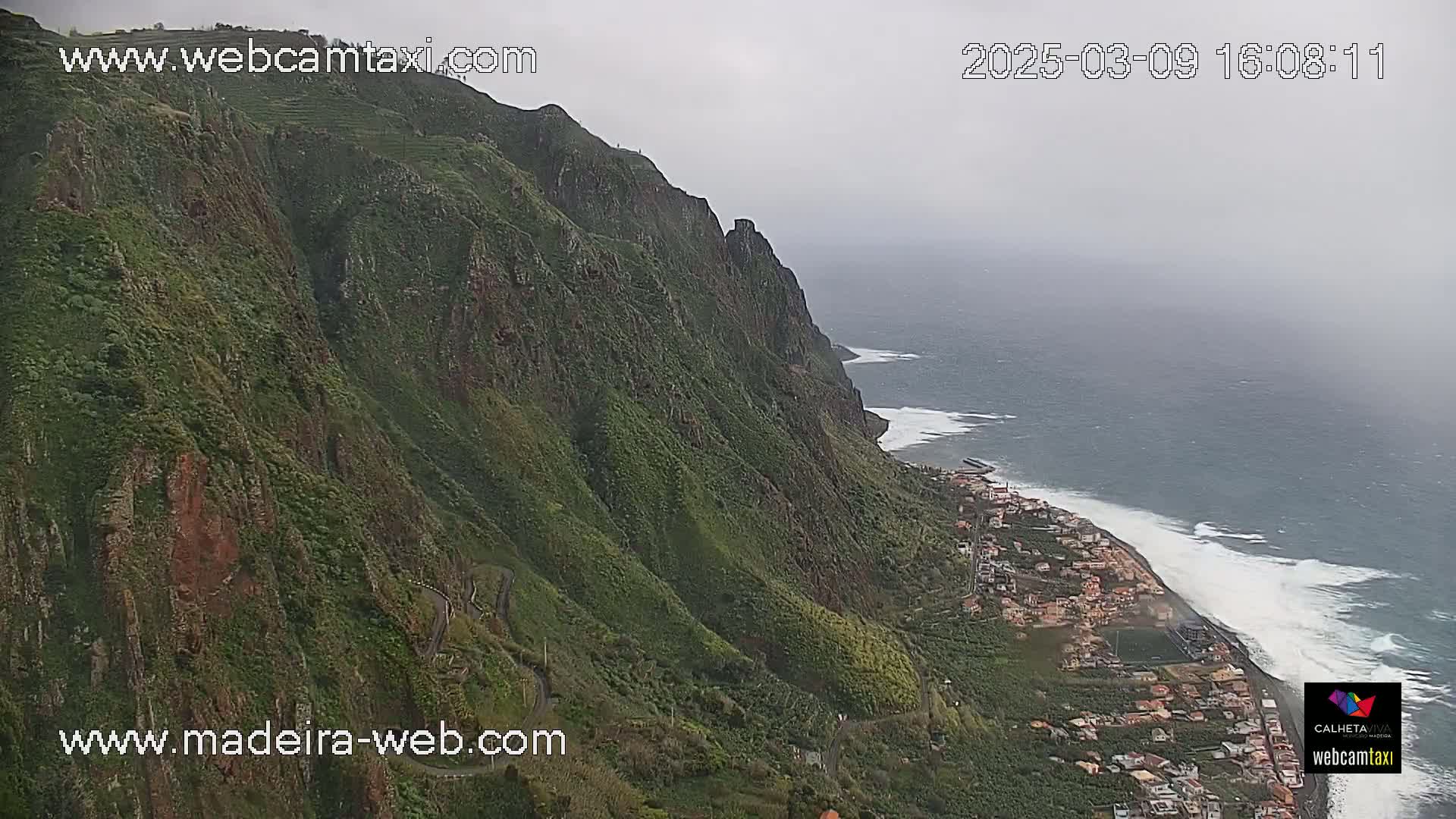 A coastal town nestled below a steep, verdant cliffside, with a grey, overcast sky and a choppy ocean.