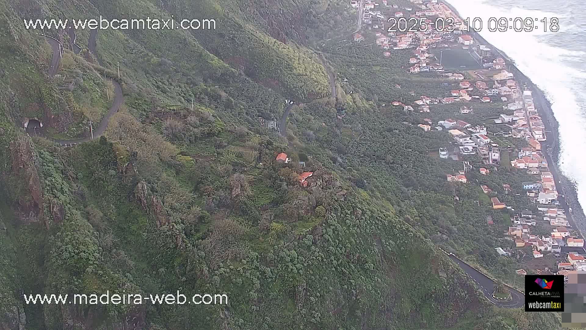 An aerial view shows a winding road descending a lush, green hillside toward a coastal village with a rocky shoreline and ocean waves.