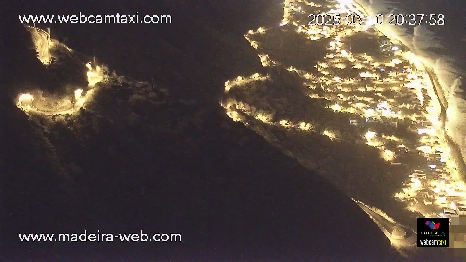 A nighttime aerial view shows a coastal town with illuminated buildings, winding roads snaking along a dark hillside, and a beach with waves visible in the distance.