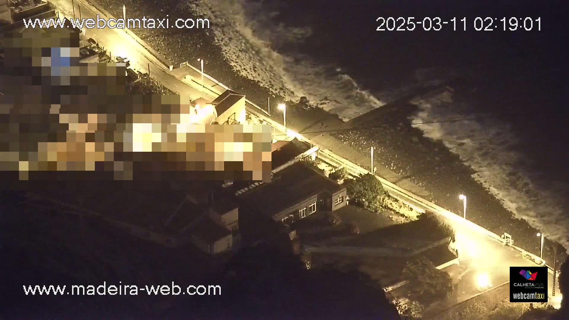 A nighttime aerial view shows a coastal road next to a rocky beach with waves crashing against the shore, and buildings illuminated by streetlights.