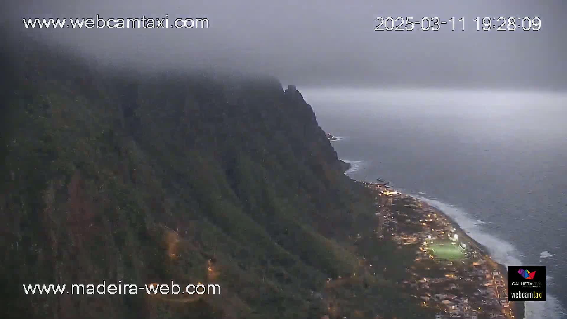 A foggy coastal scene at dusk shows a steep, dark cliff overlooking a town illuminated by lights along the ocean shore.