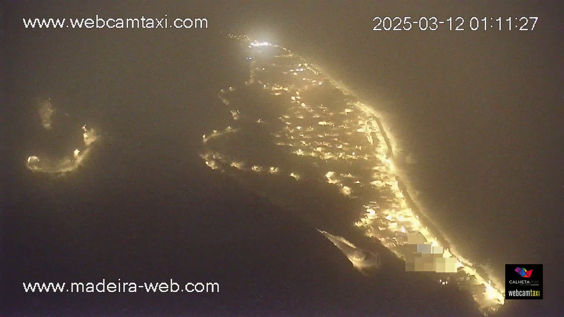 An aerial nighttime view of a coastal town, illuminated by many lights, under hazy or foggy conditions.