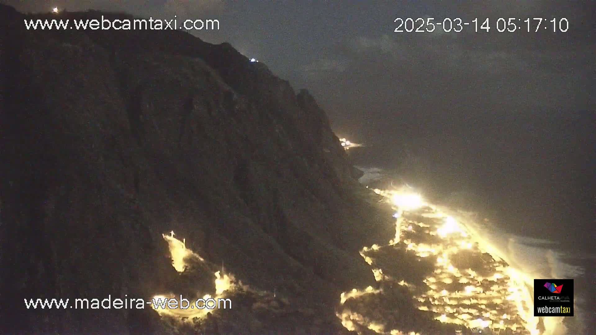 A nighttime coastal scene shows a dark cliff overlooking a town illuminated by many lights, with waves crashing on the shore under a dark sky.