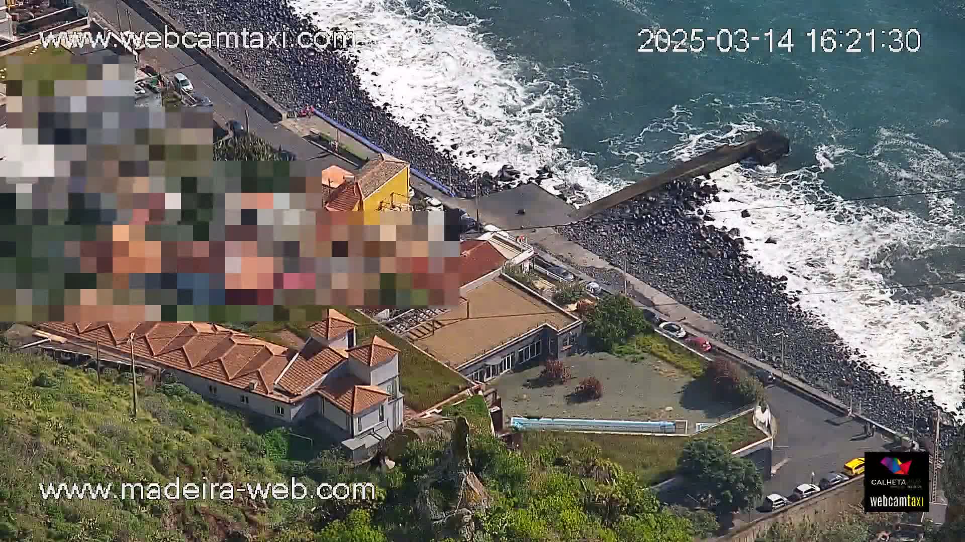 An aerial view shows a rocky coastline with crashing waves on a sunny day, alongside buildings with red tile roofs and a few cars parked along a road.