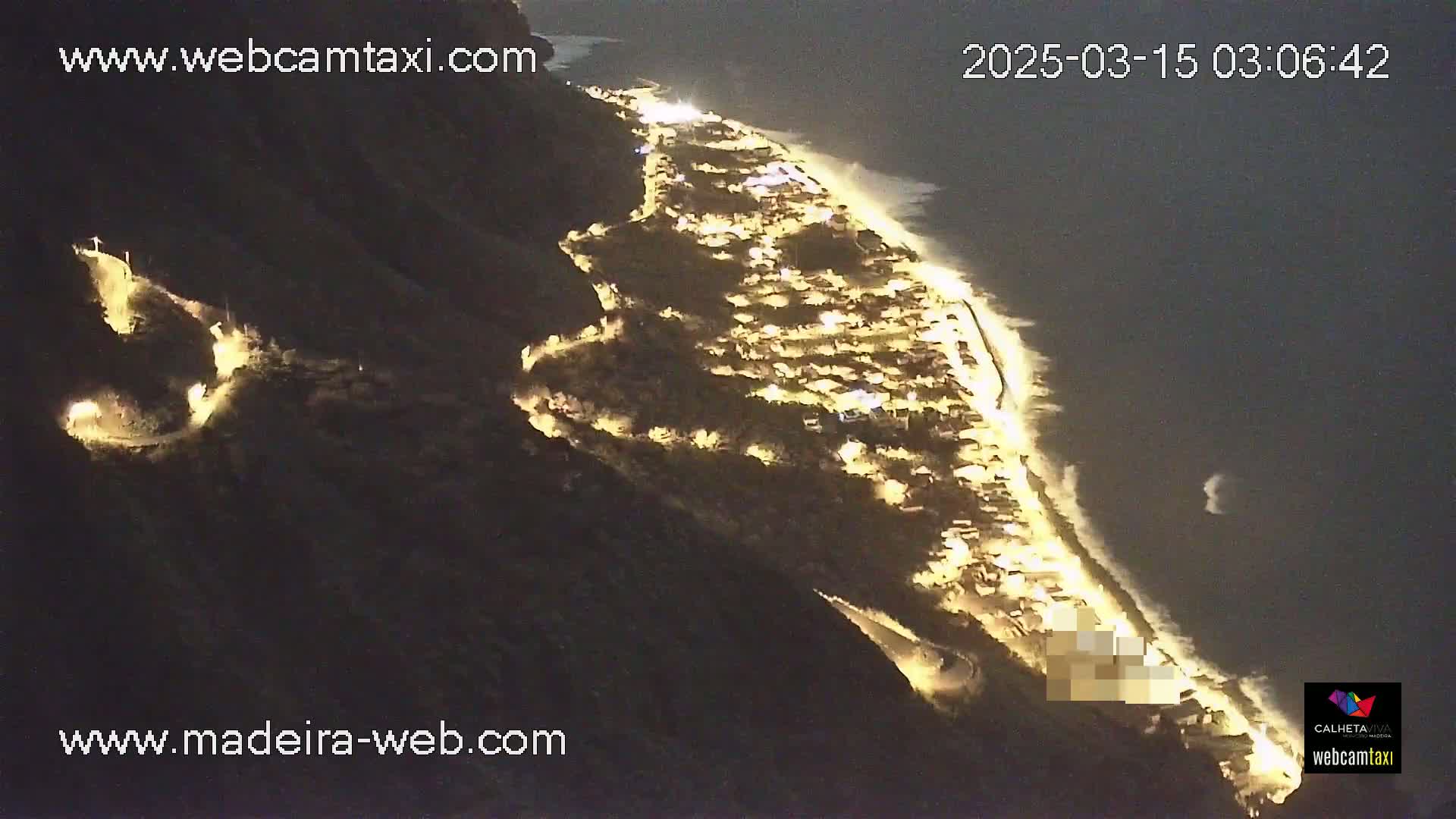 A nighttime aerial view shows a coastal town illuminated along a dark cliffside, with waves gently lapping the shore.