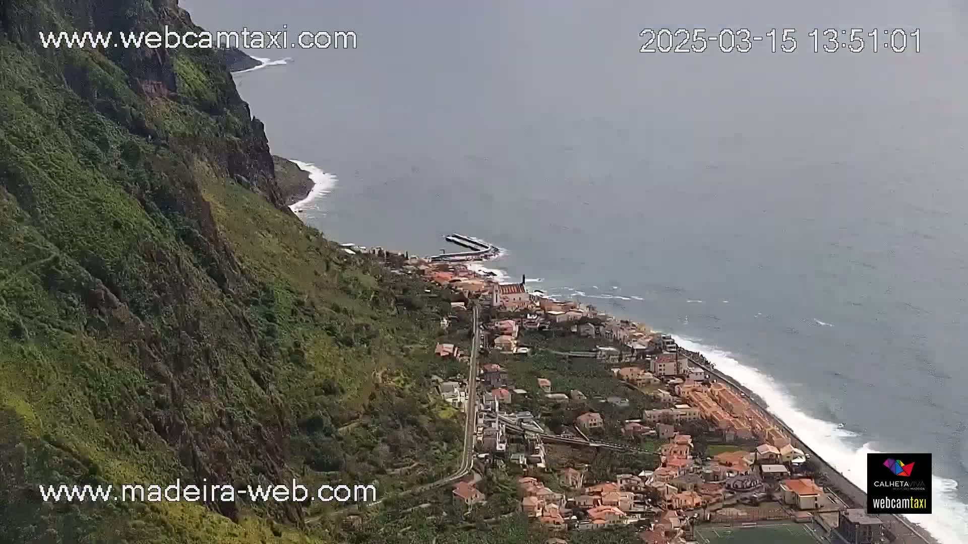 A coastal town nestled beside a steep, green cliff overlooking a gray, somewhat overcast ocean with gentle waves.