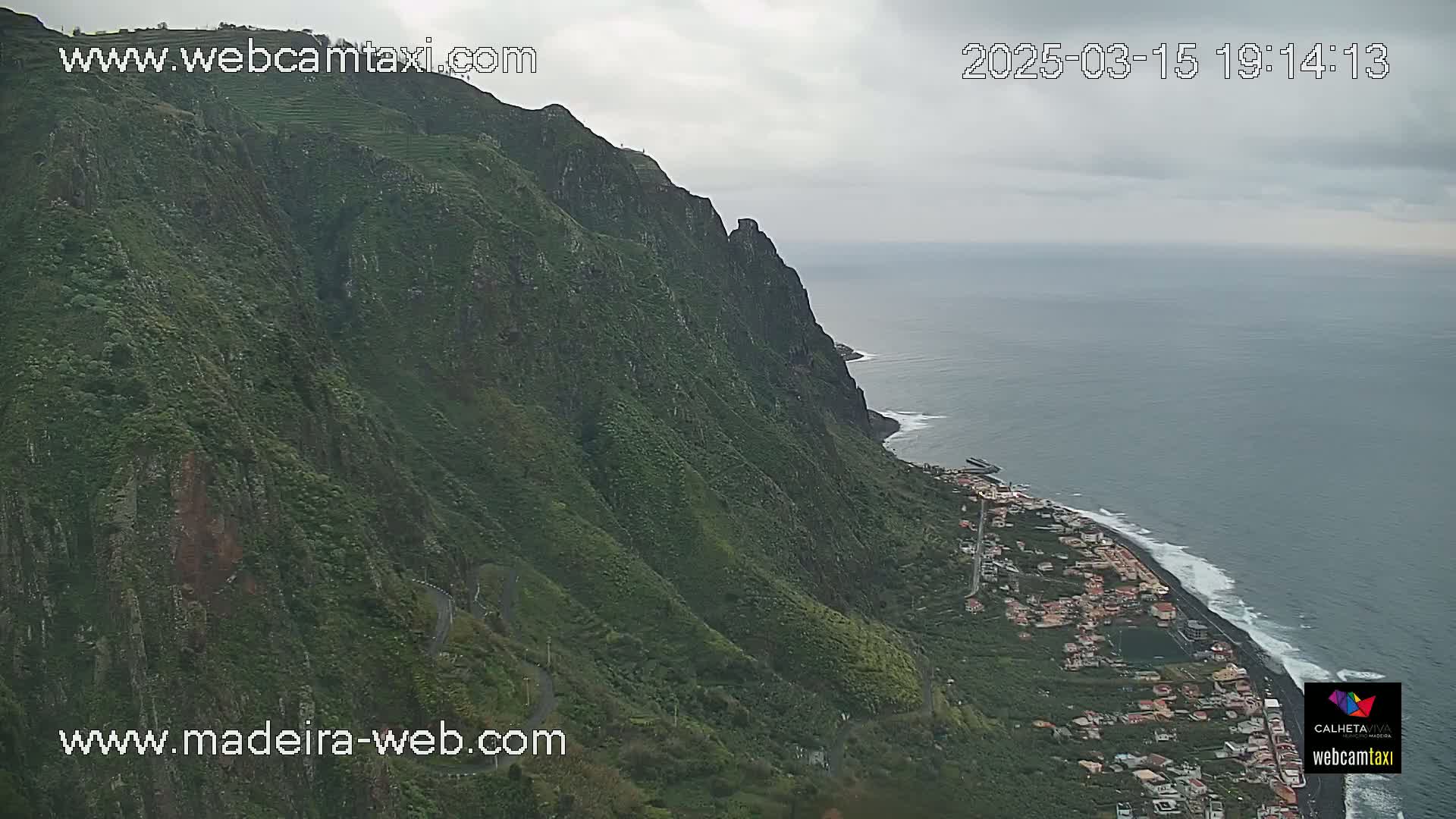 A steep, green mountainside overlooking a coastal town and ocean under overcast skies.