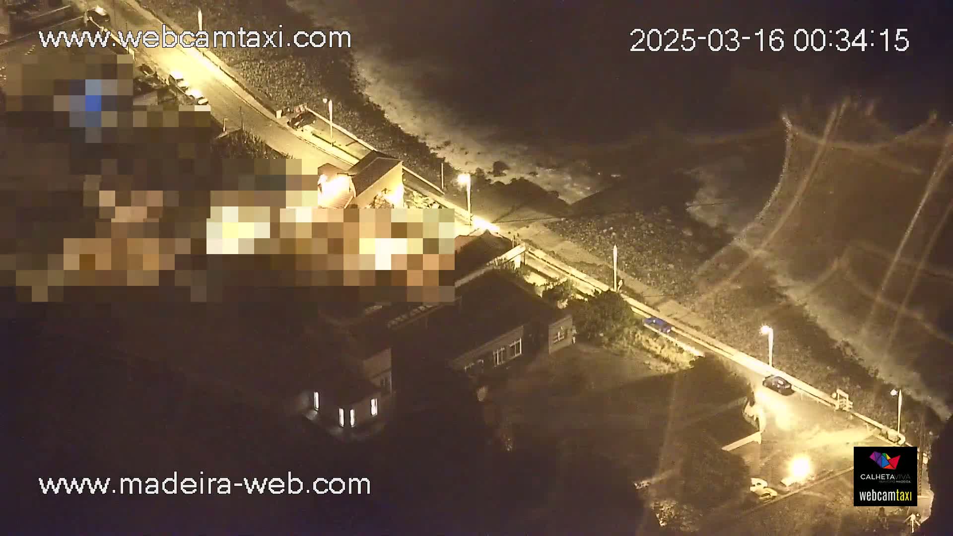 An aerial nighttime view shows a coastline with buildings and cars along a paved road, under dark, cloudy conditions.