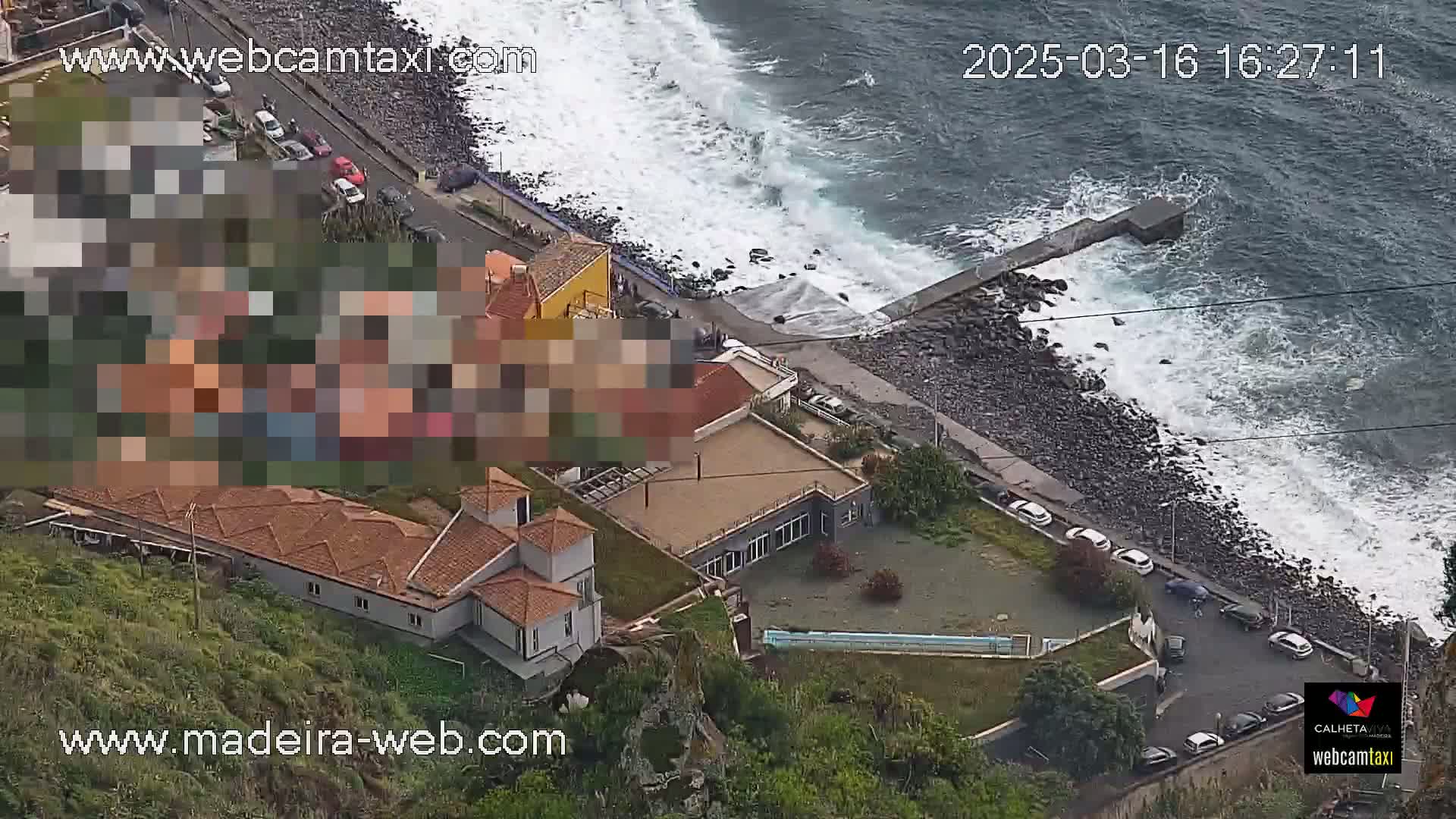 An aerial view shows a rocky coastline with crashing waves, a road with parked cars, and several buildings on a hillside.