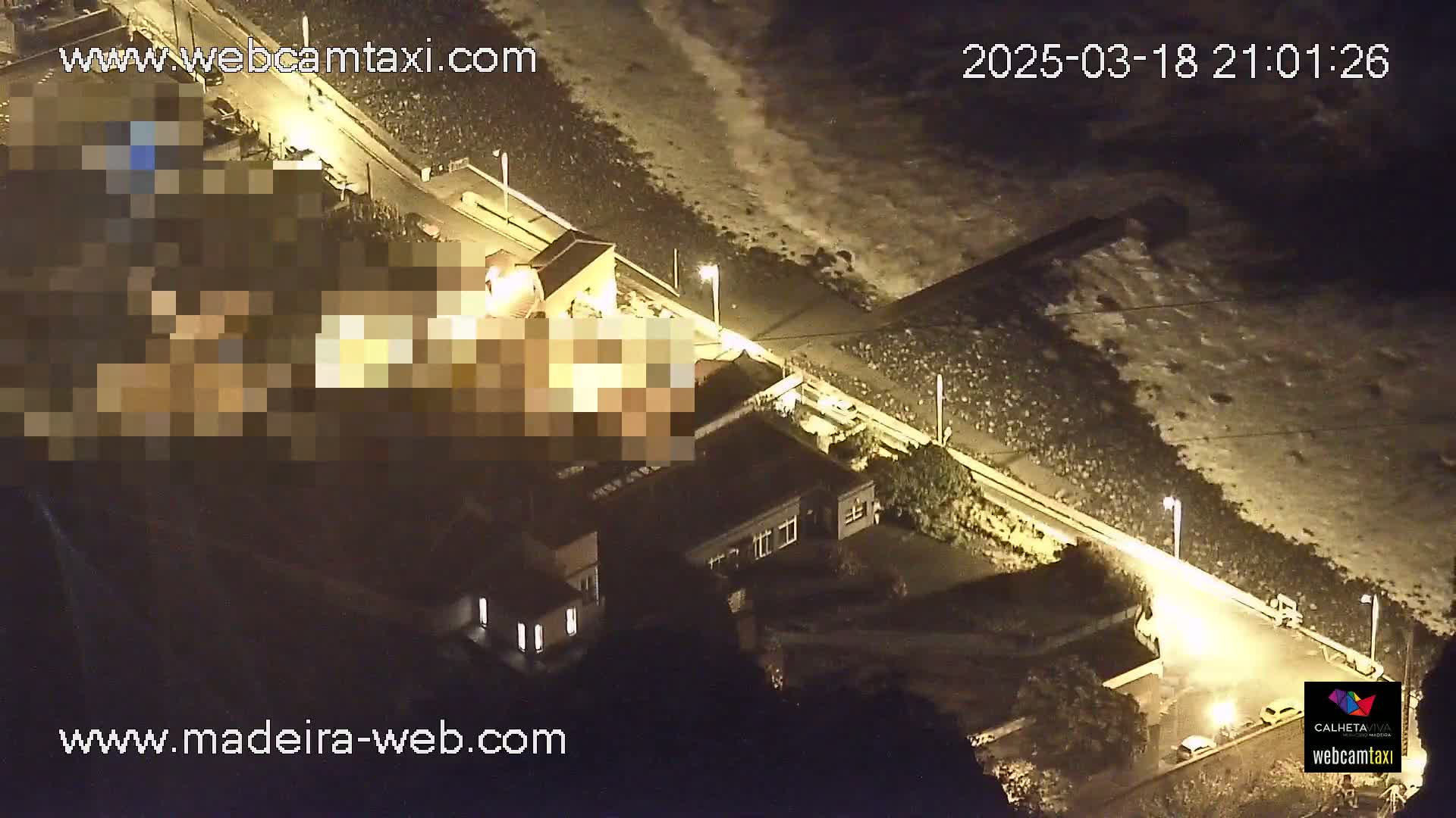 An aerial nighttime view shows a coastal road with buildings and a rocky shoreline with waves crashing against it.