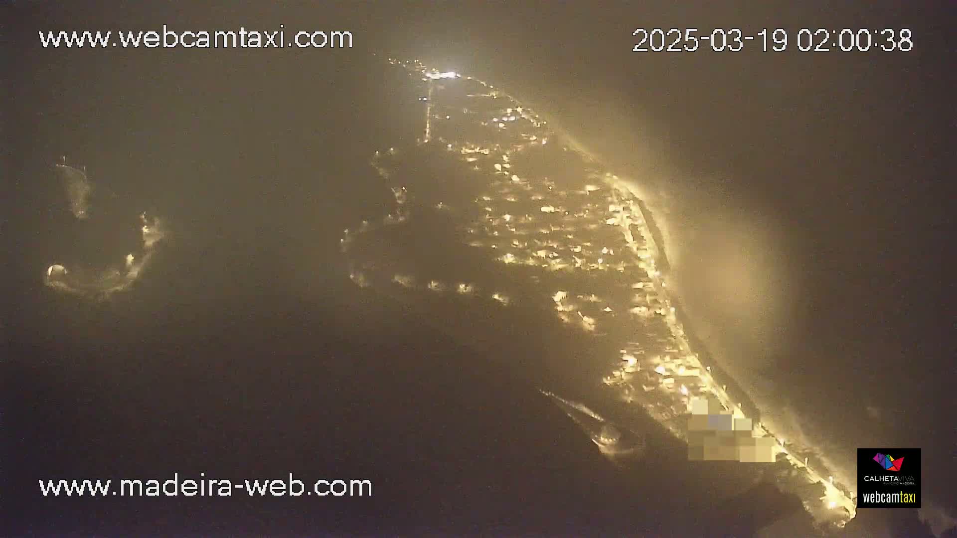 A nighttime aerial view shows a coastal town with lights illuminating the buildings and streets, partially obscured by fog or mist.