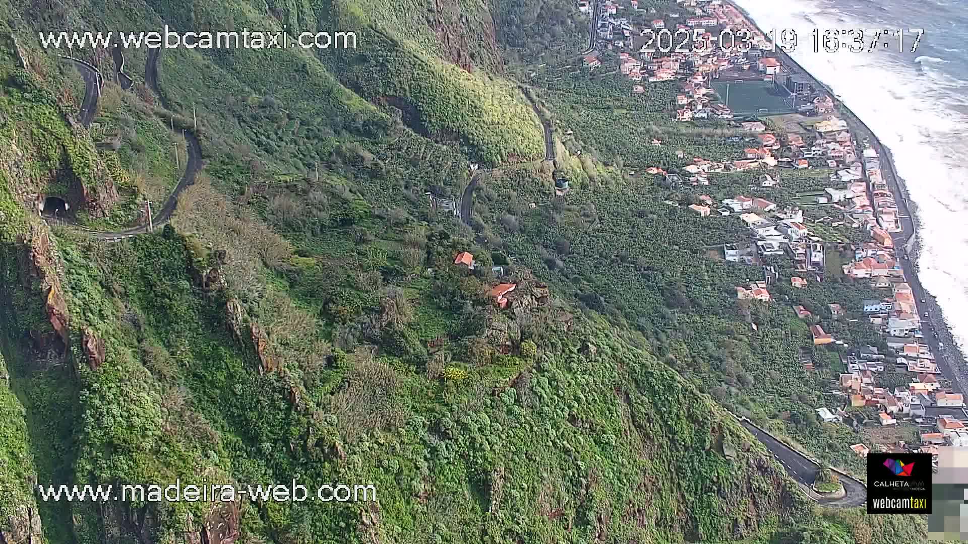 A winding road snakes down a lush, green hillside overlooking a coastal town and ocean with moderate waves.