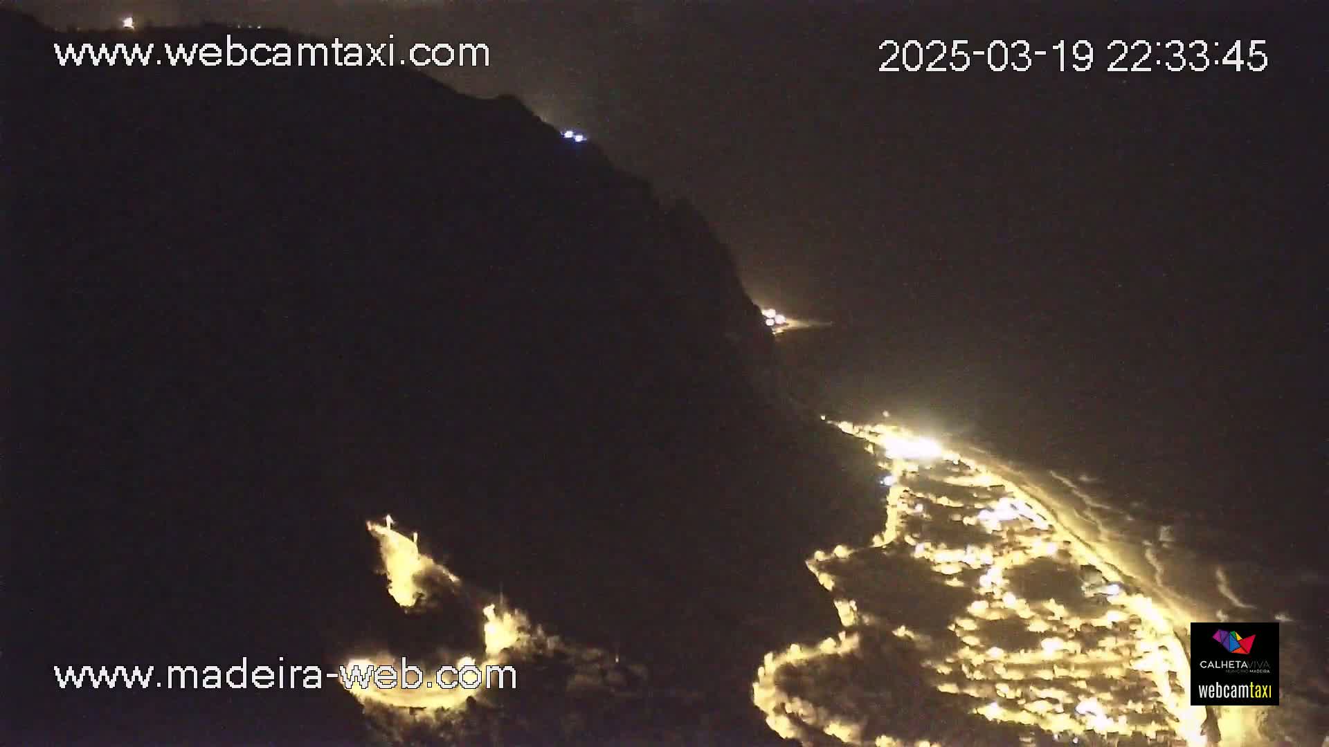 A nighttime, high-angle view shows a dark, mountainous coastline with a town illuminated below, featuring softly crashing waves.