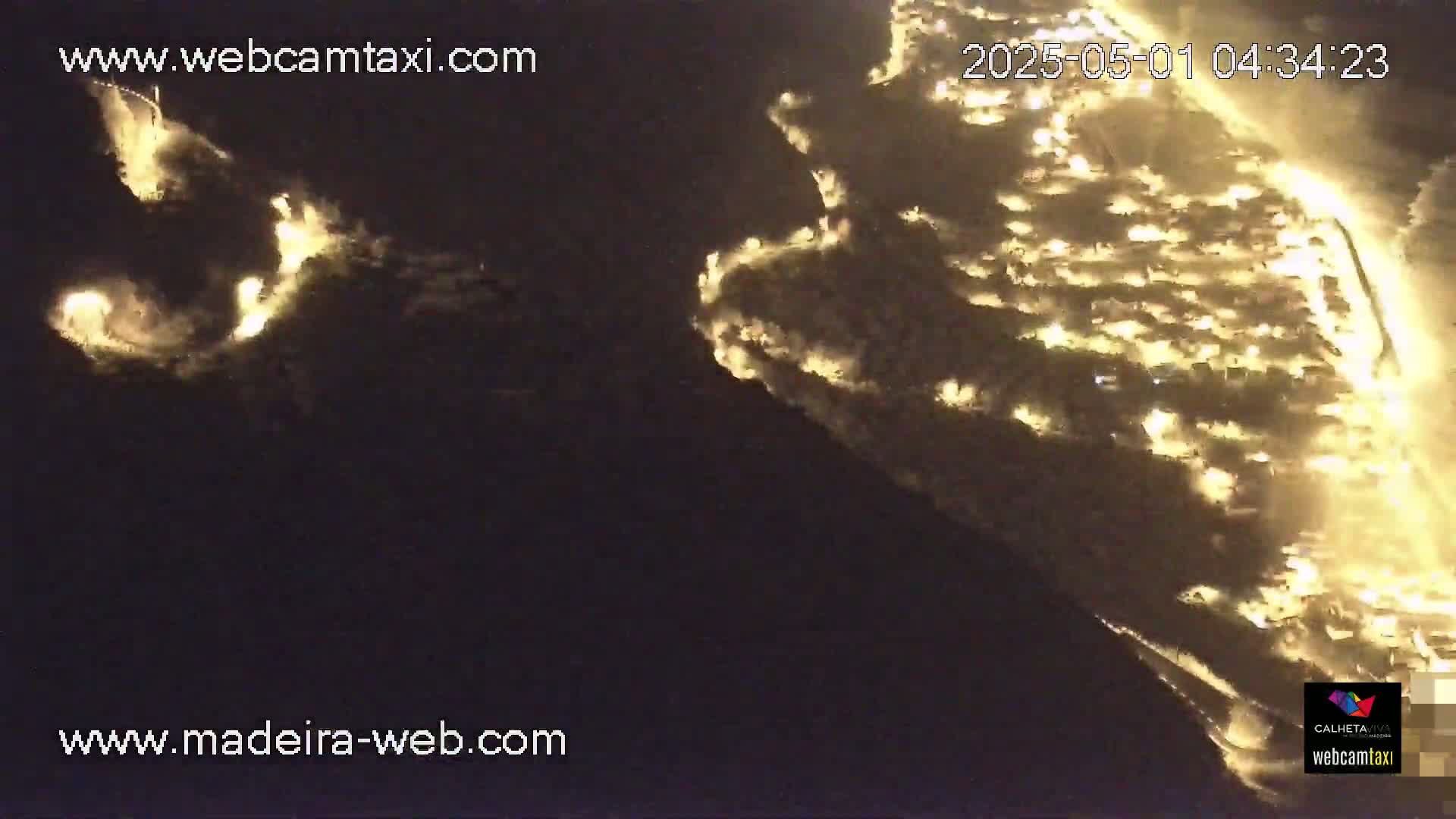 An aerial nighttime view shows a coastal town illuminated, with a fire or similar bright light source visible in the distance.