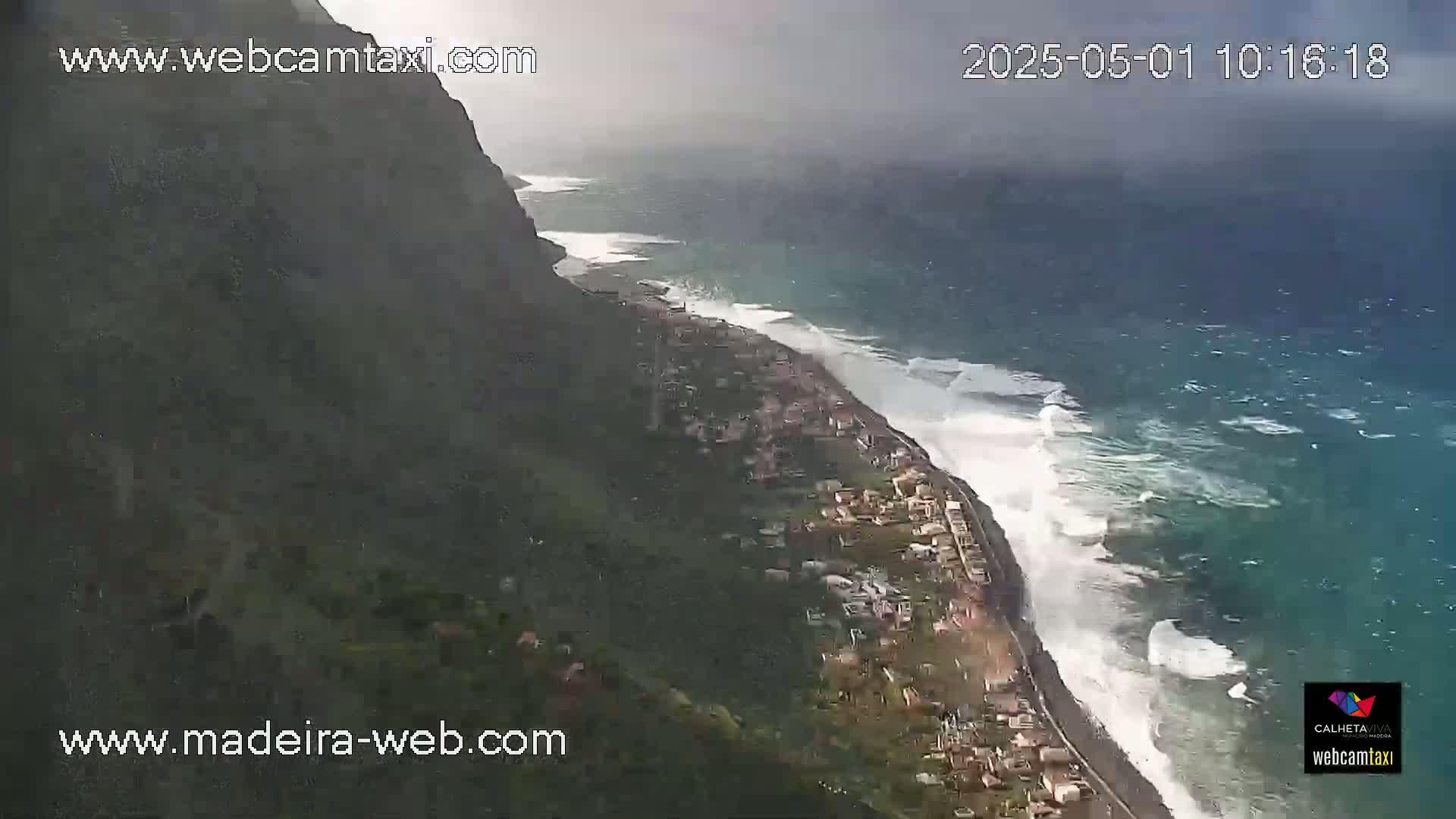A coastal town is nestled against a steep, dark cliffside overlooking a turbulent ocean under an overcast sky.