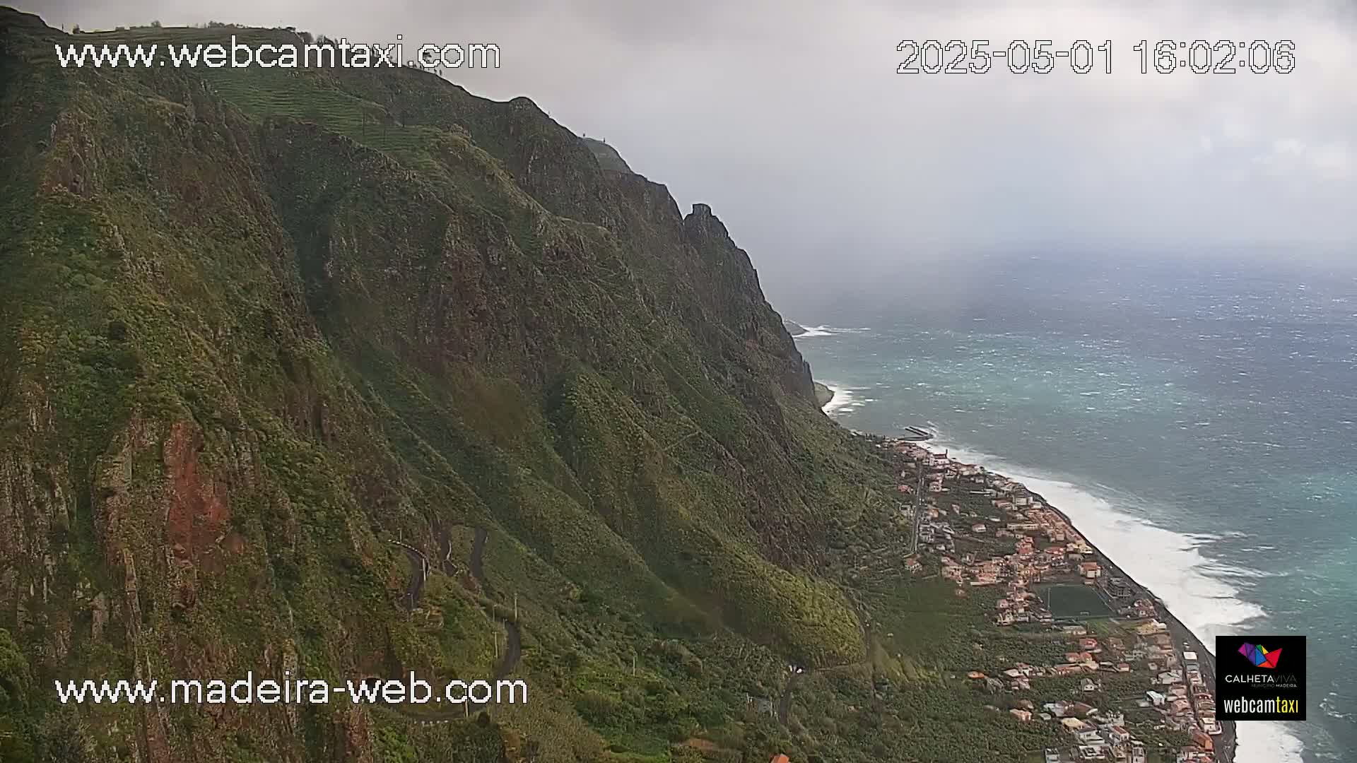 A steep, green cliffside overlooks a town situated along a wavy ocean coast under an overcast sky.