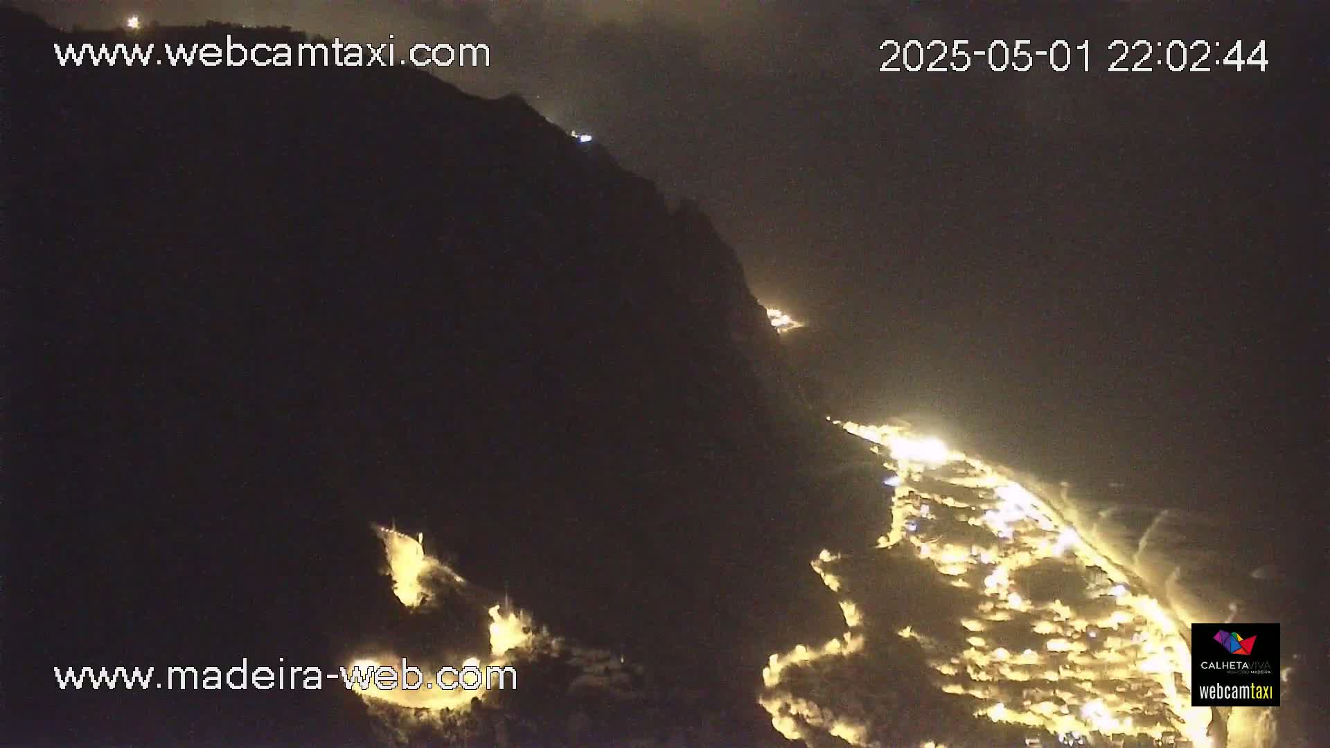 A nighttime, aerial view shows a coastal town illuminated below a dark, mountainous cliff.