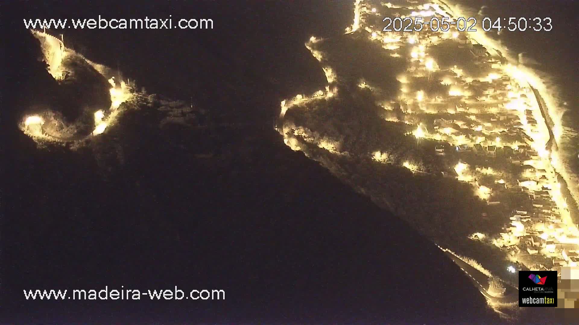 An aerial nighttime view shows a coastal town illuminated by lights, with roads winding along the dark hillsides.