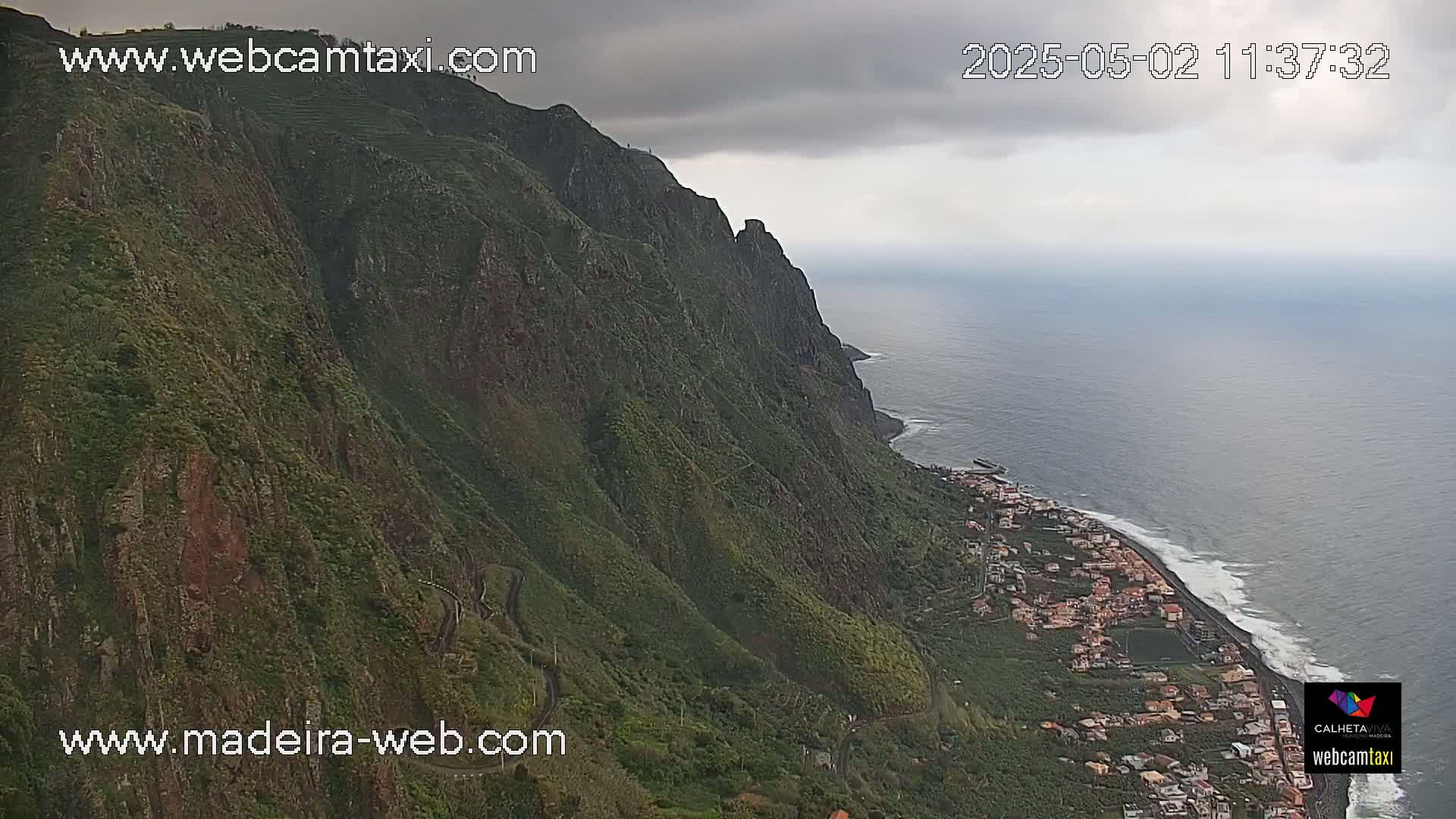 A steep, green cliffside overlooks a coastal town and ocean under an overcast sky.