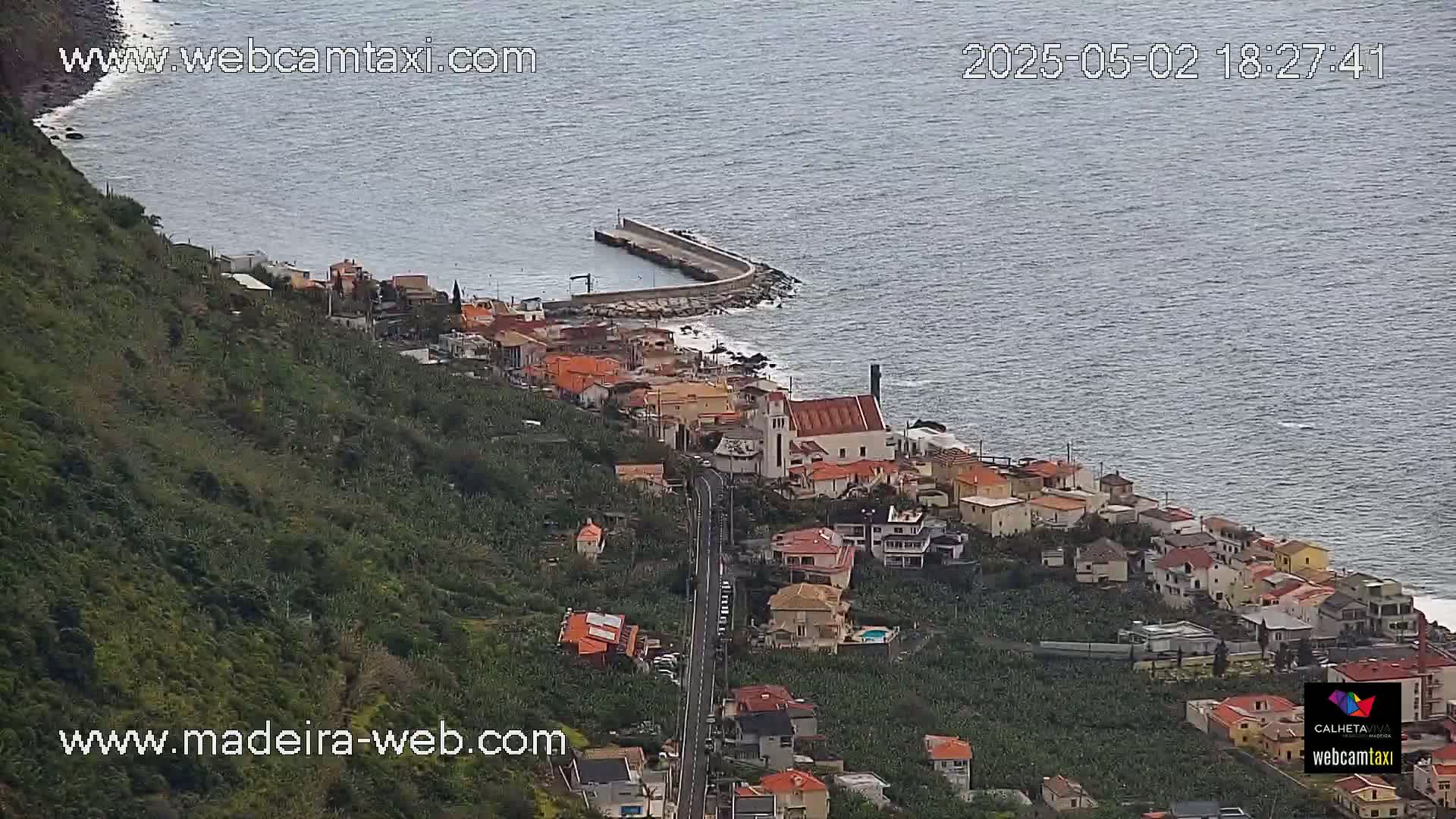 A coastal town with red-roofed houses clings to a hillside overlooking a calm ocean, a small harbor visible in the foreground.