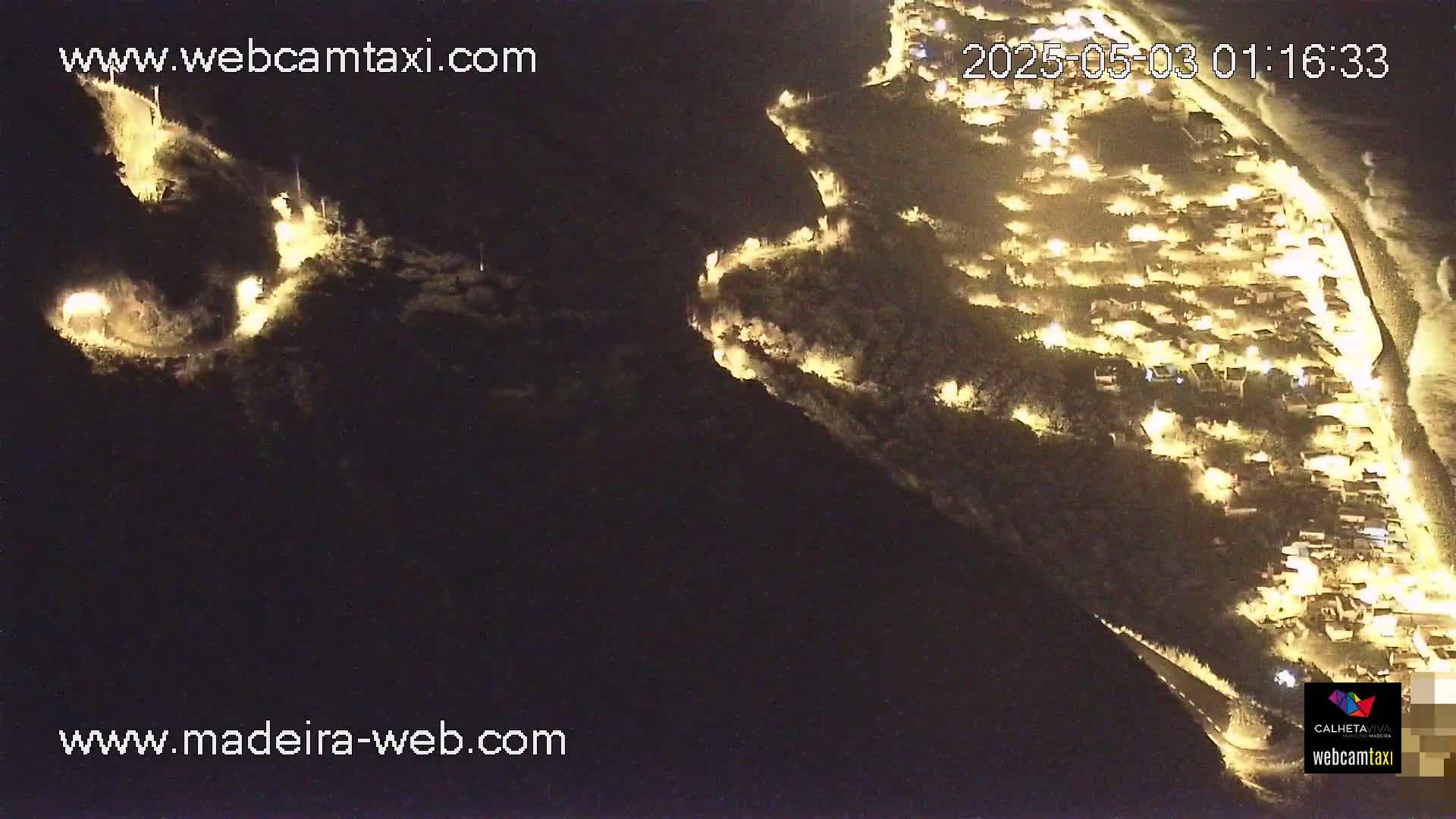 An aerial nighttime view shows a coastal town illuminated by lights, nestled between dark hillsides.