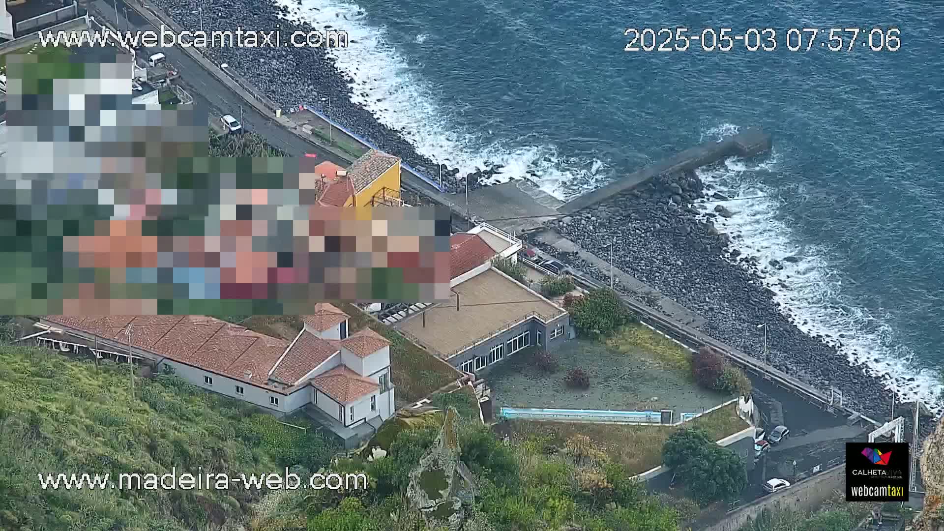 An aerial view shows several buildings near a rocky coastline with moderate waves on a partly cloudy day.