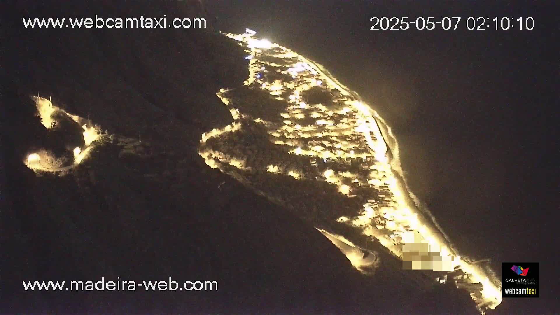 An aerial nighttime view shows a coastal town illuminated, nestled on a cliffside next to a dark ocean.