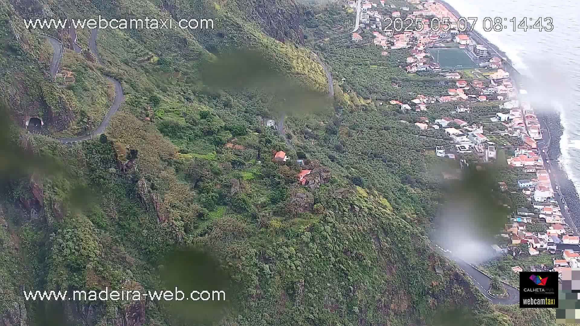 An aerial view shows a winding road curving down a lush green hillside to a coastal town with a rocky beach and overcast conditions.
