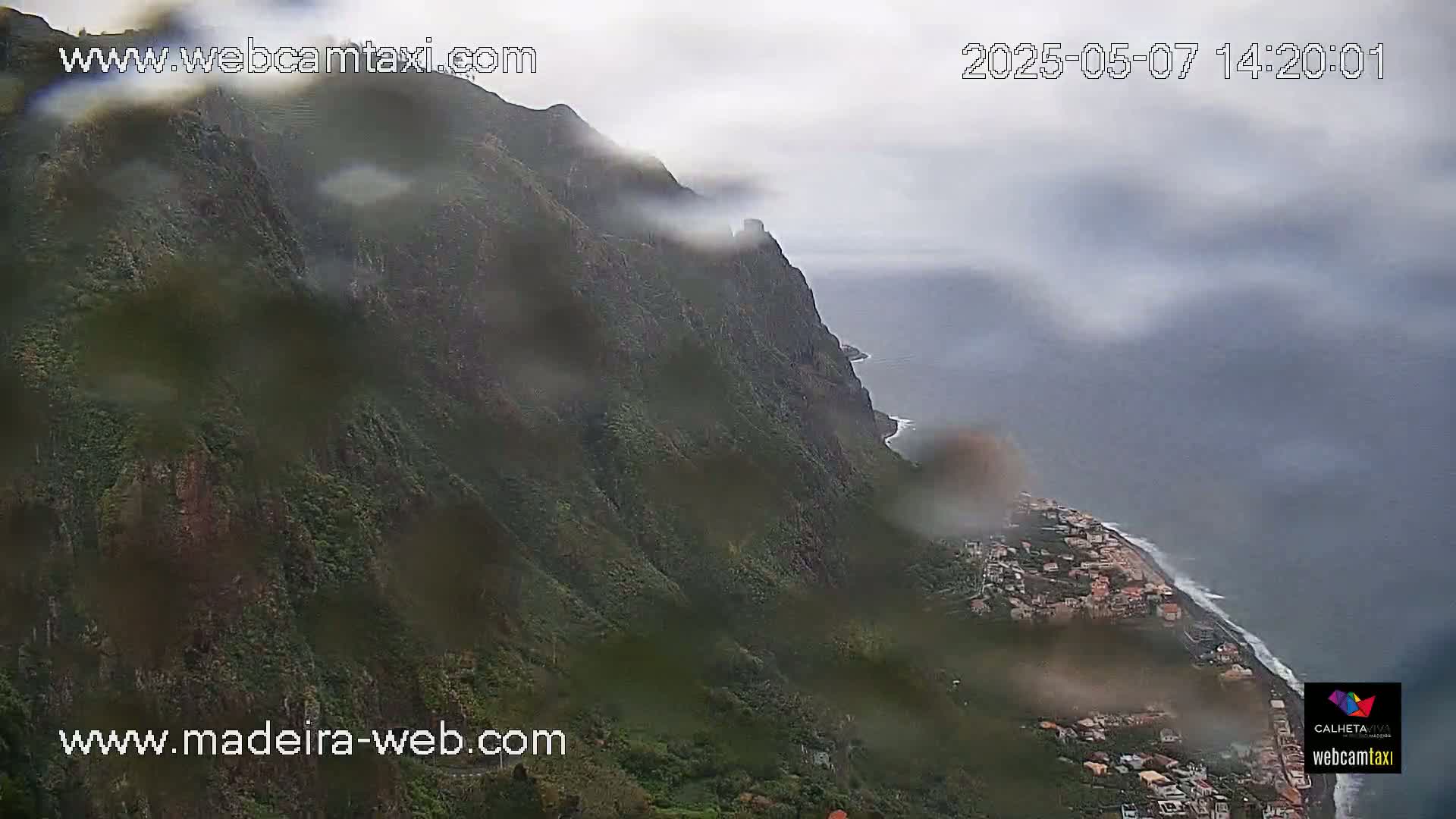 A rain-covered coastal cliffside reveals a town nestled below along the ocean, under an overcast sky.